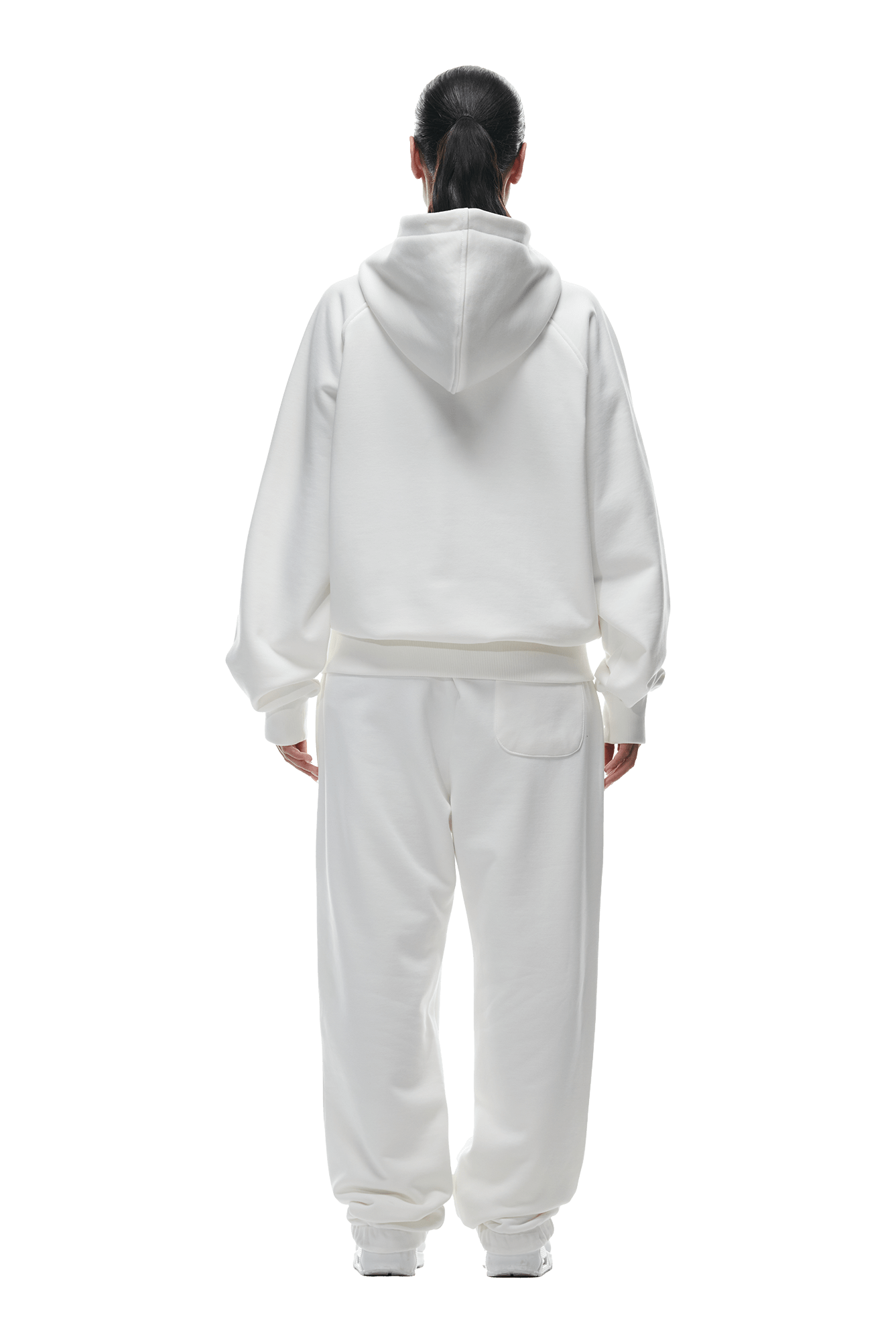 A person with long dark hair in a ponytail stands facing away, wearing the 6PM ZIP HOODIE COCONUT WHITE with matching white sweatpants. The clean finish makes this outfit ideal for everyday wear. Background is transparent.