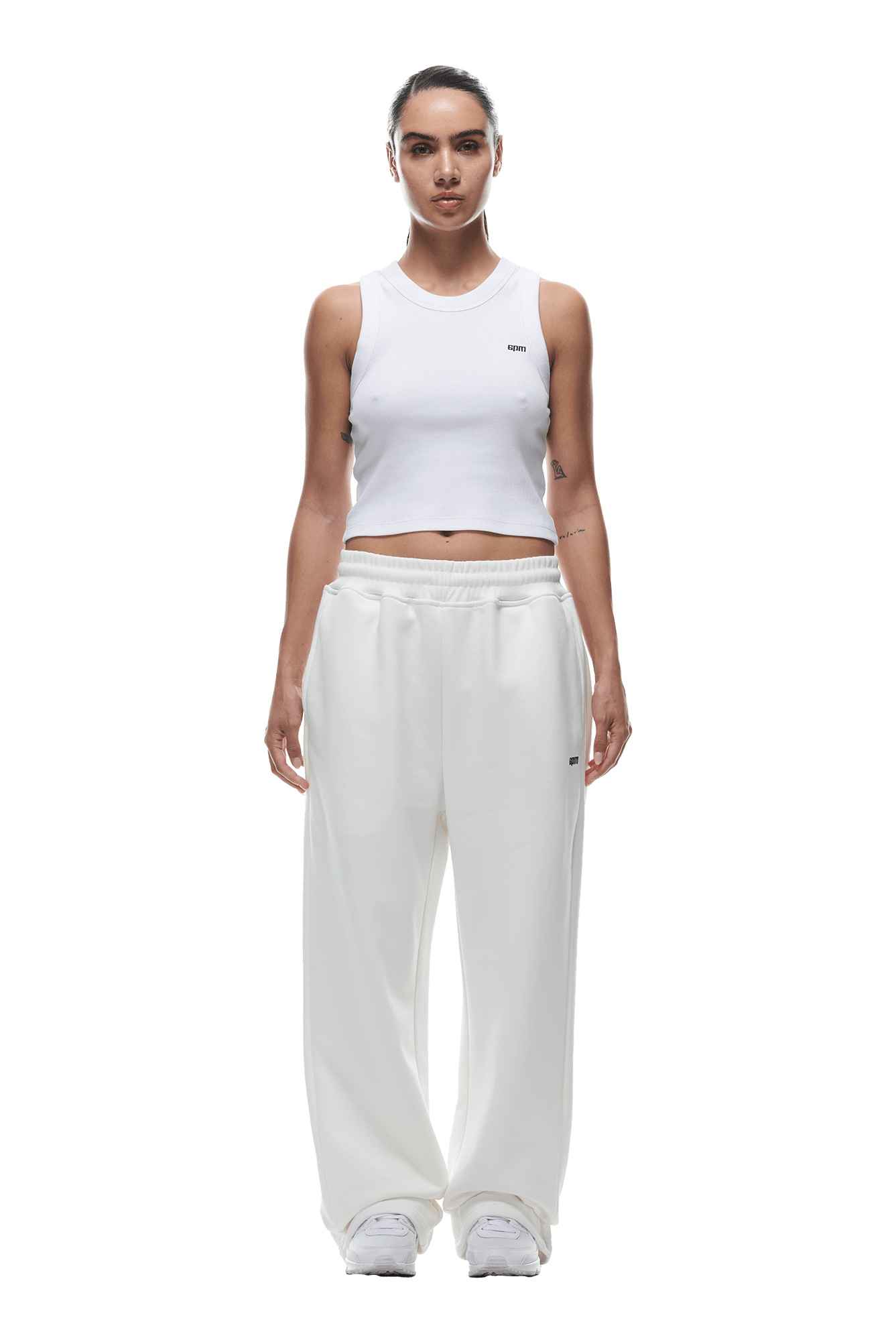 A woman faces forward wearing the 6PM OPEN LEG JOGGER COCONUT WHITE and a white sleeveless crop top, paired with white sneakers. Her dark hair is pulled back and her arms rest at her sides against a transparent background.