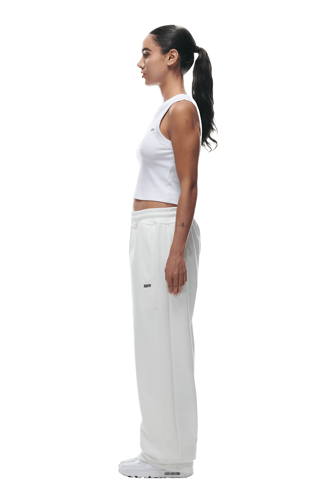 A woman in profile stands against a white background, wearing 6PMs OPEN LEG JOGGER COCONUT WHITE pants with a white sleeveless crop top, her dark hair in a ponytail, completed with white sneakers.
