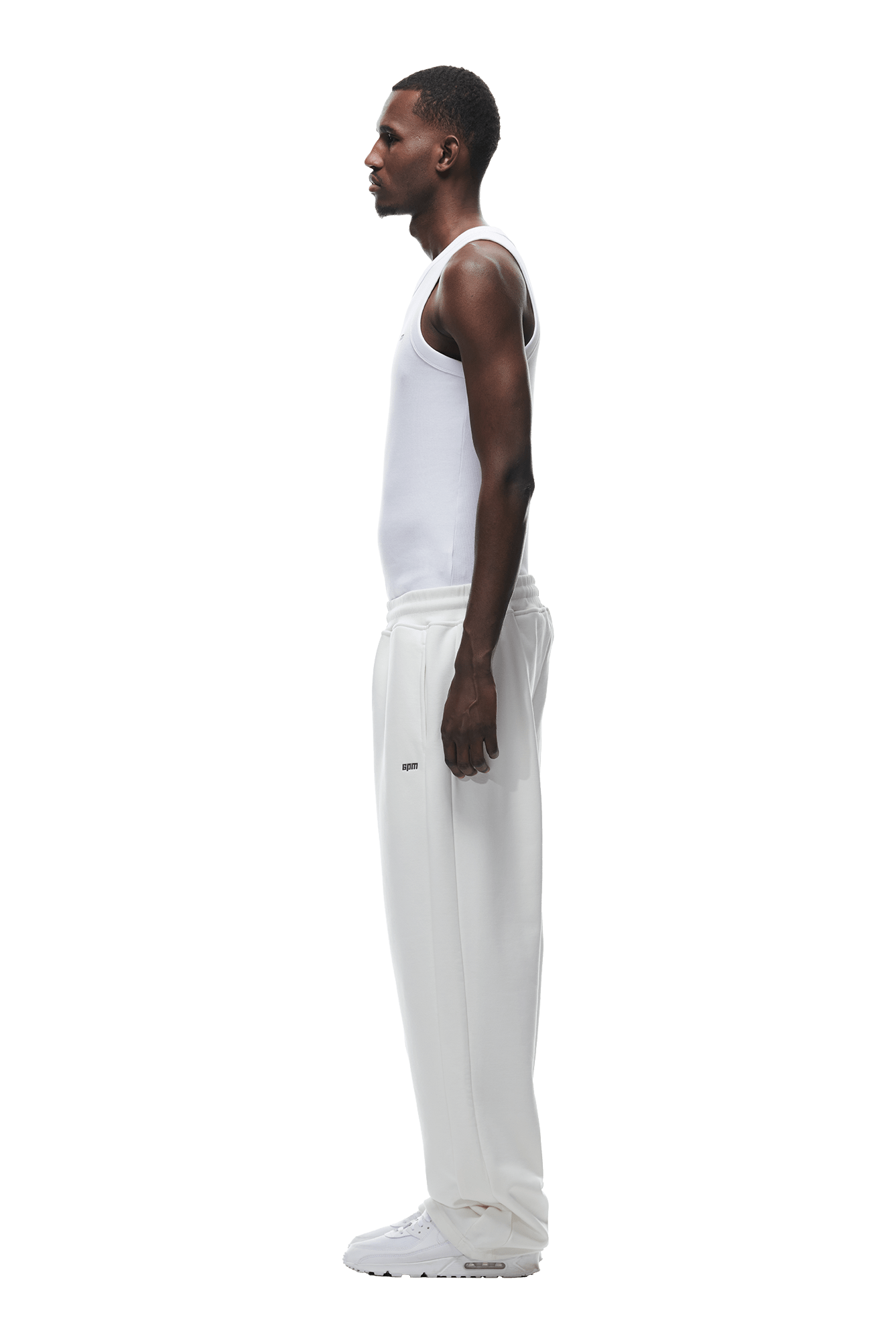 A man stands in profile facing left, wearing a white tank top, 6PM OPEN LEG JOGGER COCONUT WHITE sweatpants with a modern stacked fit, and white sneakers against a plain background.