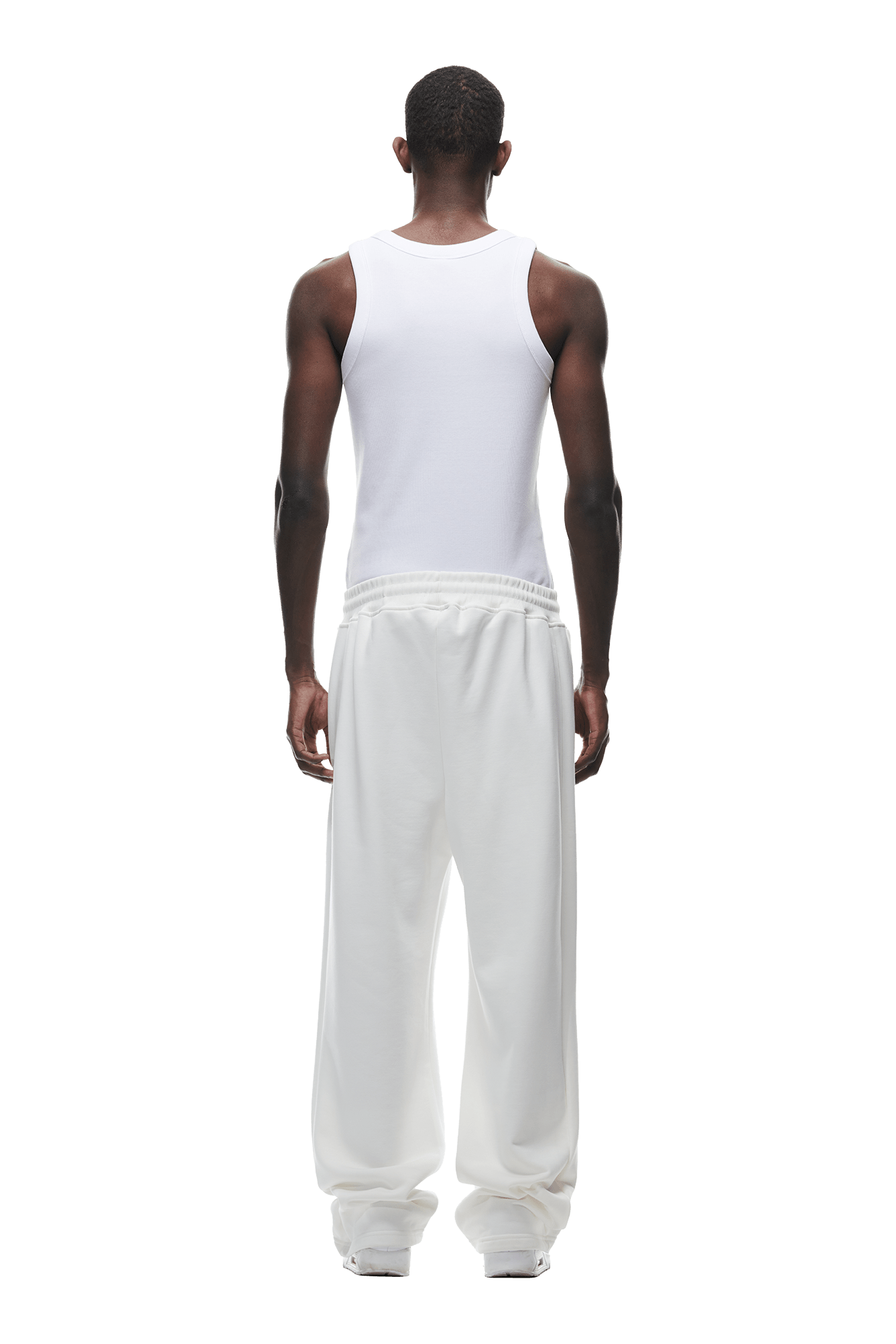 A man stands facing away, wearing a white sleeveless shirt and 6PMs OPEN LEG JOGGER COCONUT WHITE pants with a stacking length. His arms rest by his sides against a plain green background.