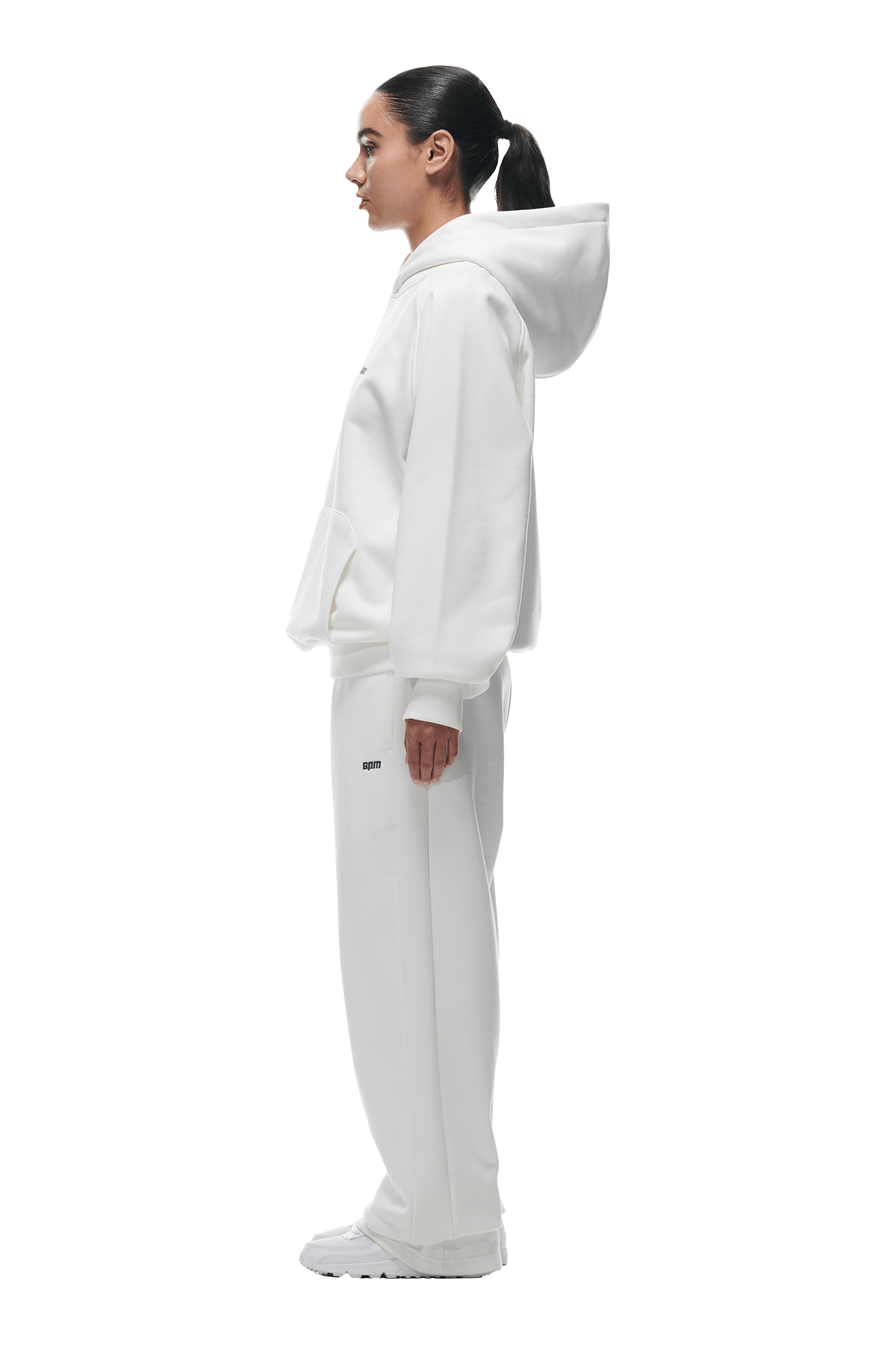 A woman with dark hair in a ponytail stands in profile, wearing the 6PM HOODIE COCONUT WHITE with a kangaroo pocket and matching sweatpants with sneakers, perfect for everyday use, against a plain background.