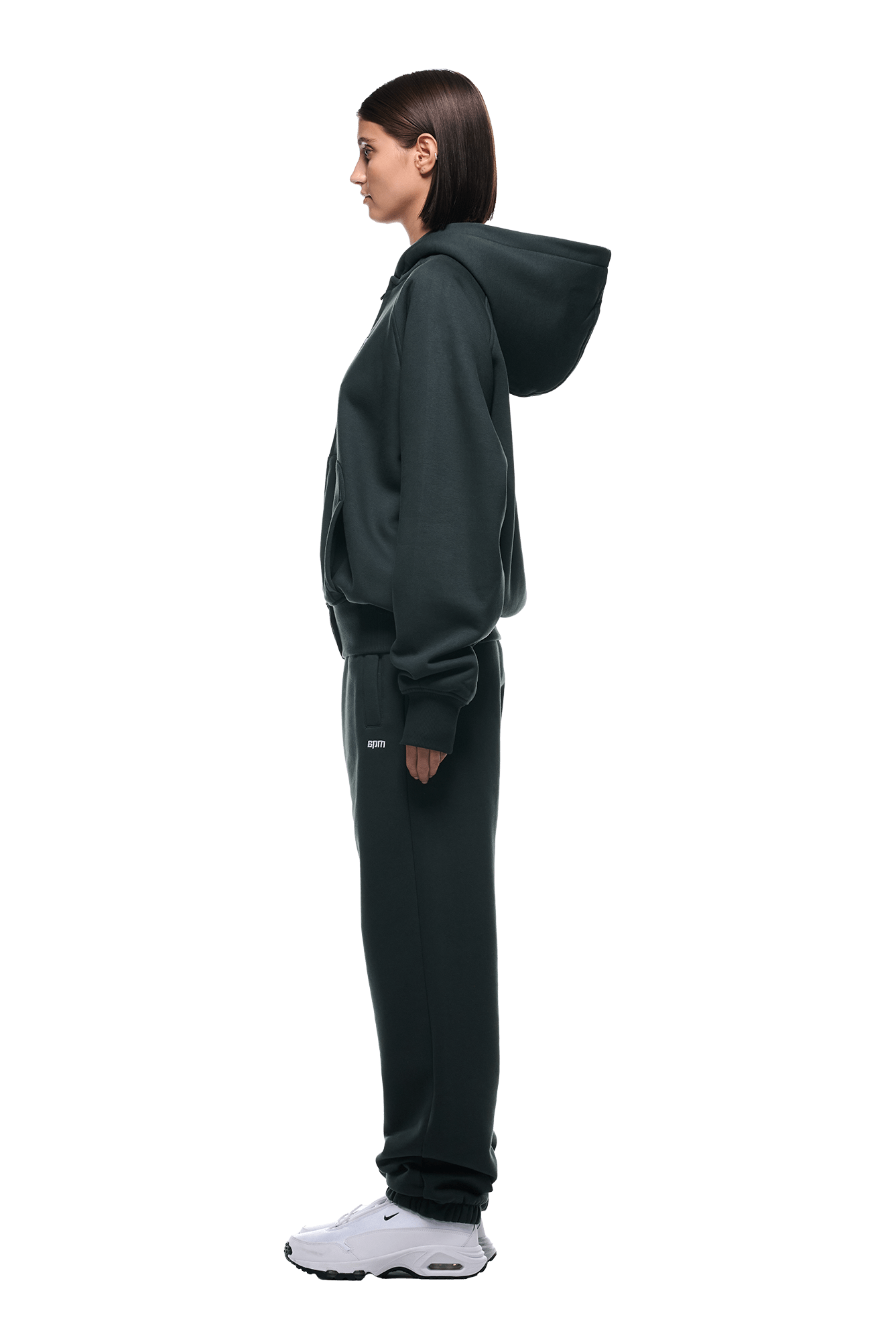 Wearing the 6PM ZIP HOODIE DARK EMERALD with matching sweatpants and white sneakers, a woman with straight, shoulder-length hair stands in profile against a plain background, showcasing the outfit’s structured style.