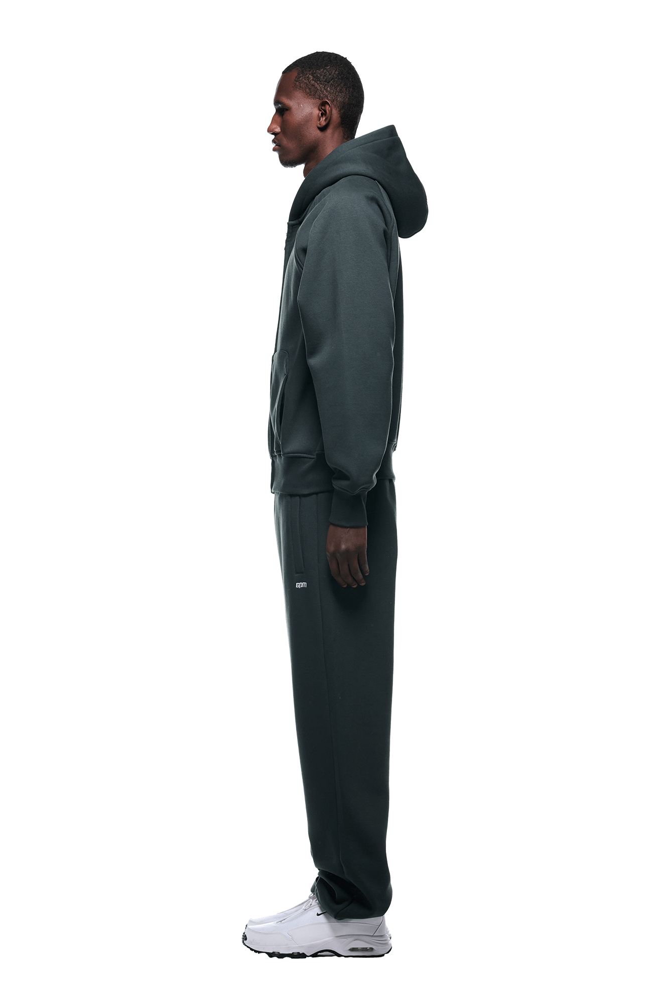 A man in profile models the 6PM ZIP HOODIE DARK EMERALD with matching sweatpants and white sneakers against a plain green backdrop, showcasing its clean finish ideal for everyday wear.