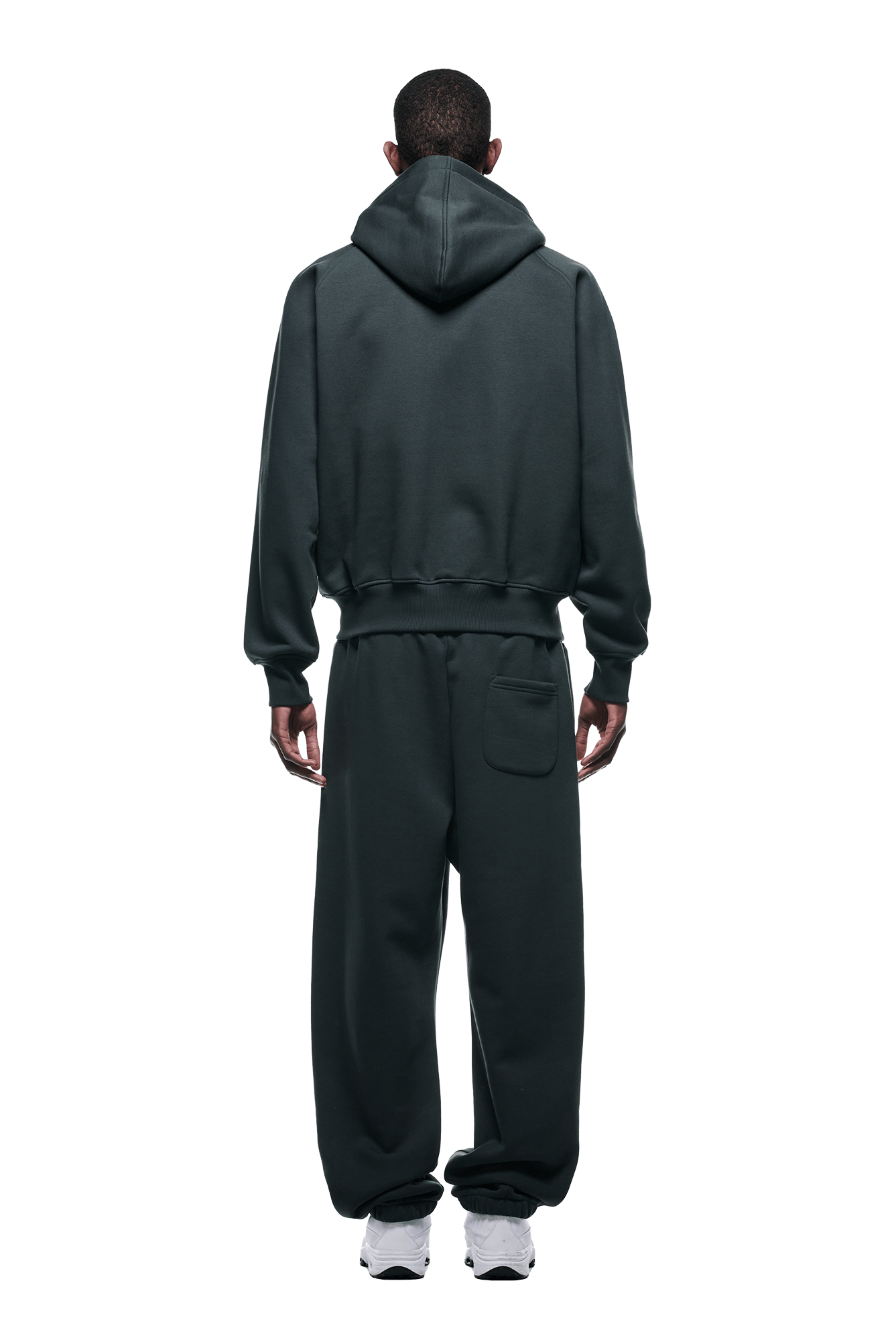A person faces away, wearing the 6PM ZIP HOODIE DARK EMERALD with hood up and matching sweatpants, featuring a back pocket. Paired with white sneakers. Clean finish, ideal for daily wear. Transparent background.