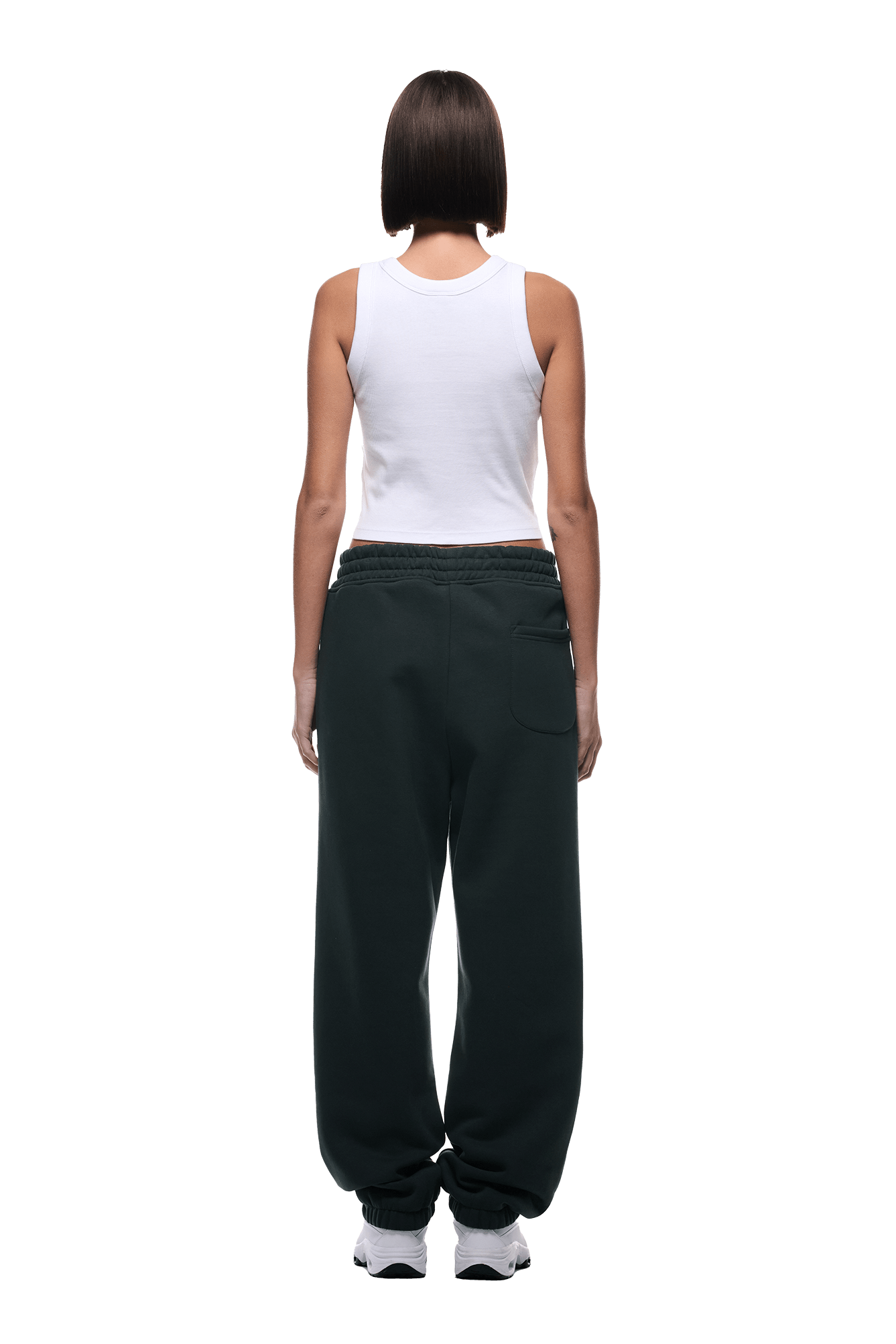 A person with short dark hair faces away, wearing a white tank top, 6PM CUFF JOGGER DARK EMERALD sweatpants with cuffed legs, and white sneakers against a plain green background.