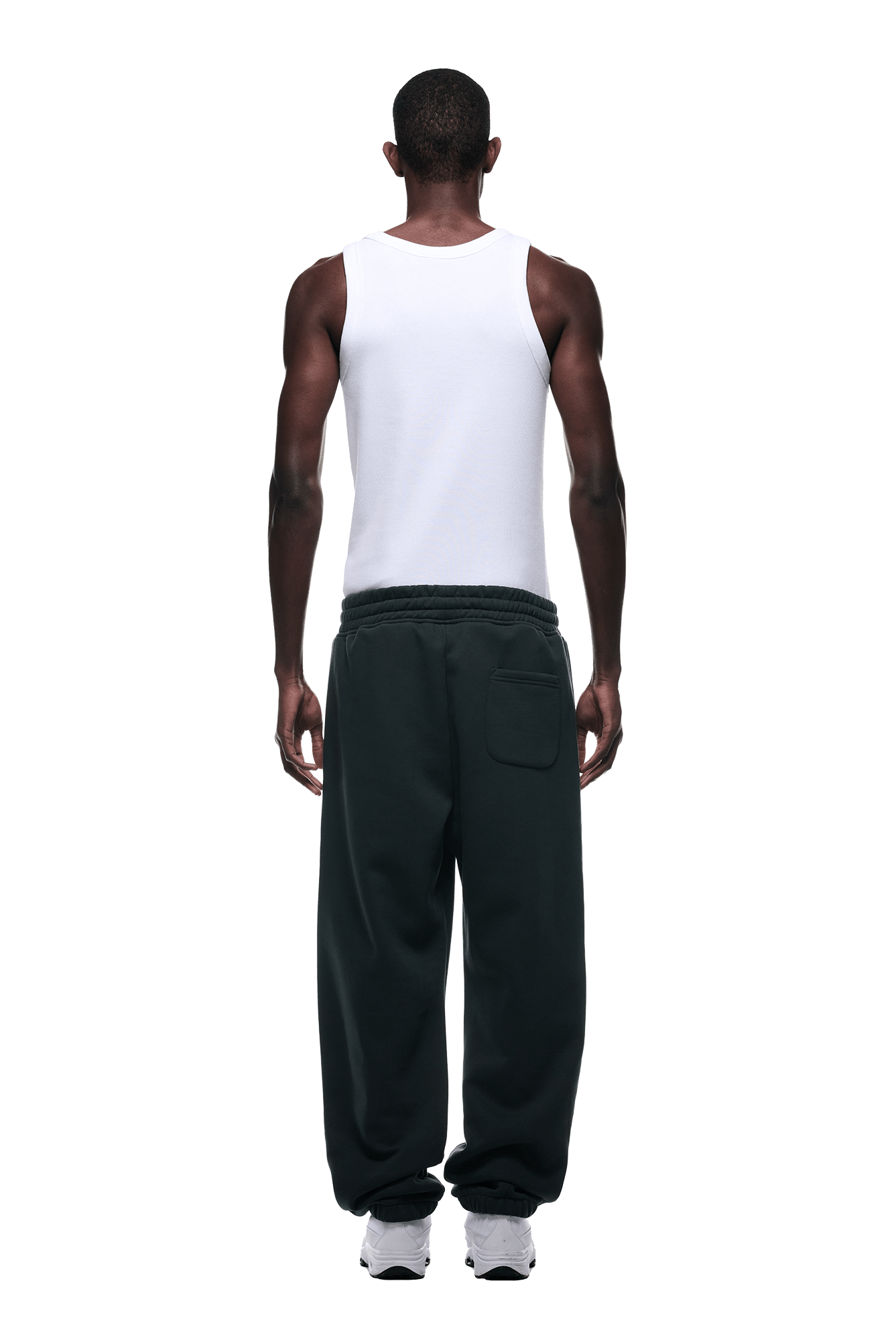 A man stands facing away, wearing a white tank top, 6PMs CUFF JOGGER DARK EMERALD sweatpants with cuffed leg openings, and white sneakers against a plain background.