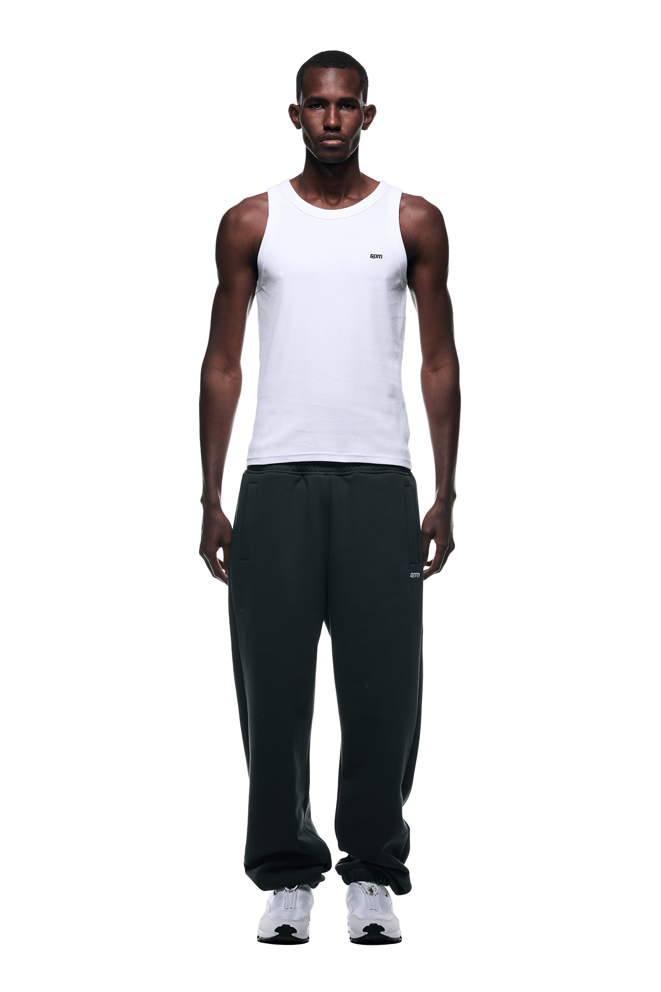 A man stands facing forward against a plain green background, wearing a white sleeveless tank top, 6PM CUFF JOGGER DARK EMERALD sweatpants with cuffed leg openings, and white sneakers.