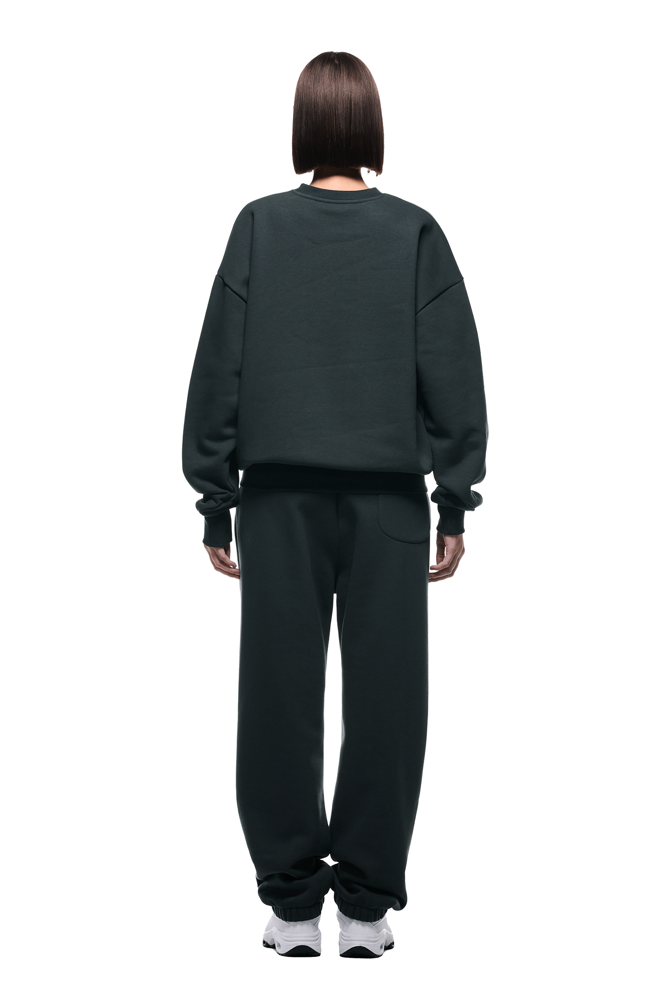 A person with straight, chin-length brown hair faces away, wearing the 6PM CREWNECK DARK EMERALD oversized sweatshirt with ribbed trims and matching sweatpants, paired with white sneakers, against a plain background.