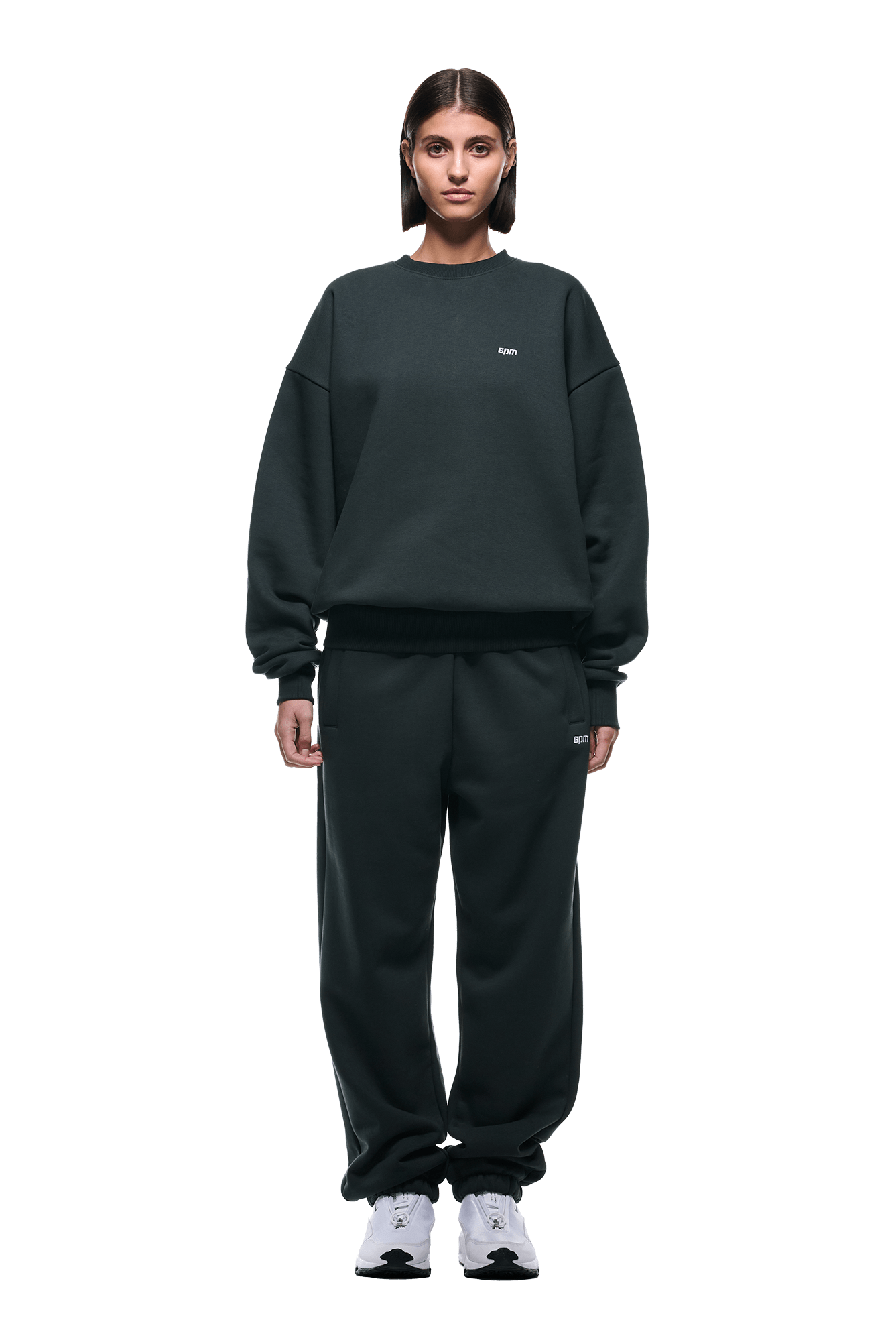 A person stands facing forward, wearing the 6PM CREWNECK DARK EMERALD sweatshirt and matching sweatpants with ribbed trims and white sneakers, set against a plain green background.