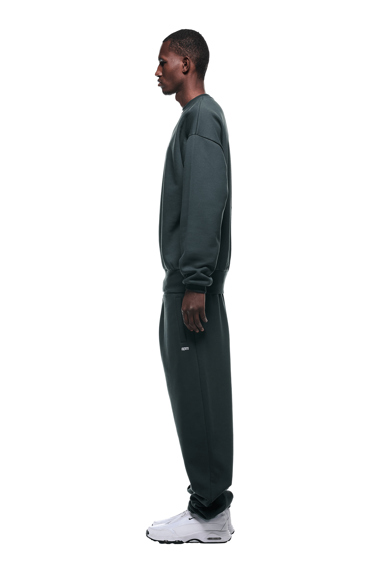 A person in profile wears the 6PM CREWNECK DARK EMERALD sweatshirt with matching sweatpants and white sneakers, isolated on a transparent background.