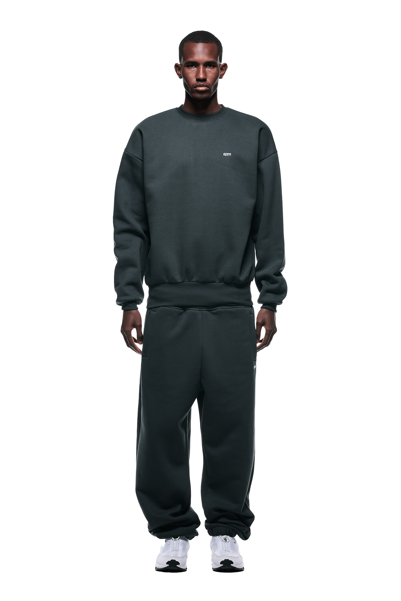 A man faces forward wearing the 6PM CREWNECK DARK EMERALD oversized sweatshirt with ribbed trims, paired with matching sweatpants and white sneakers against a solid green background.