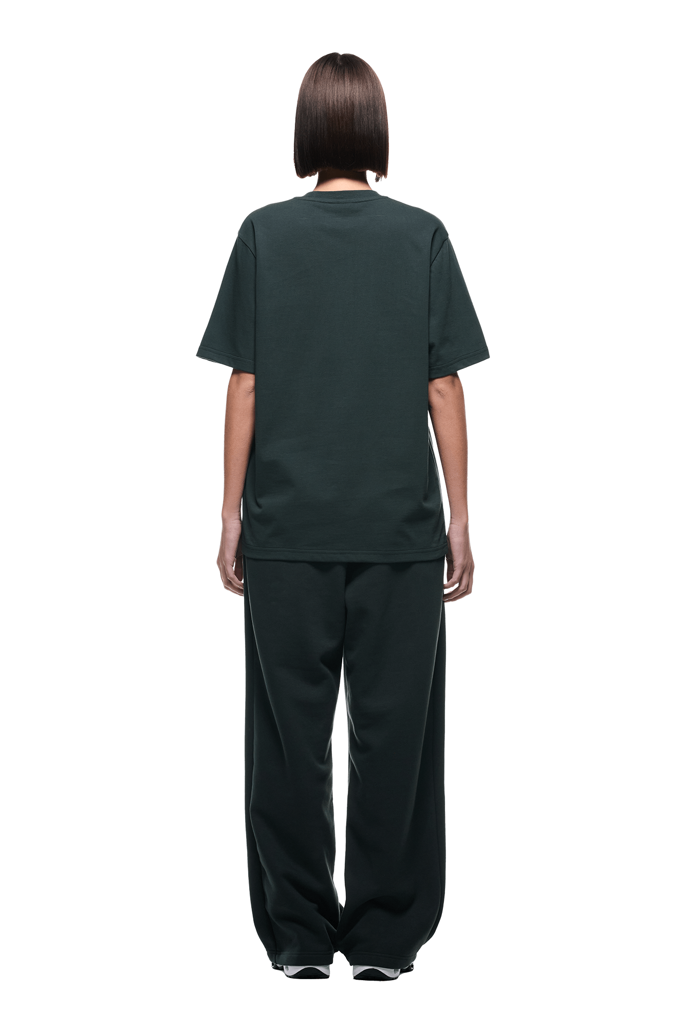 A person with short, straight brown hair faces away, wearing the 6PM T-SHIRT DARK EMERALD and matching loose pants with white sneakers, set against a plain background.