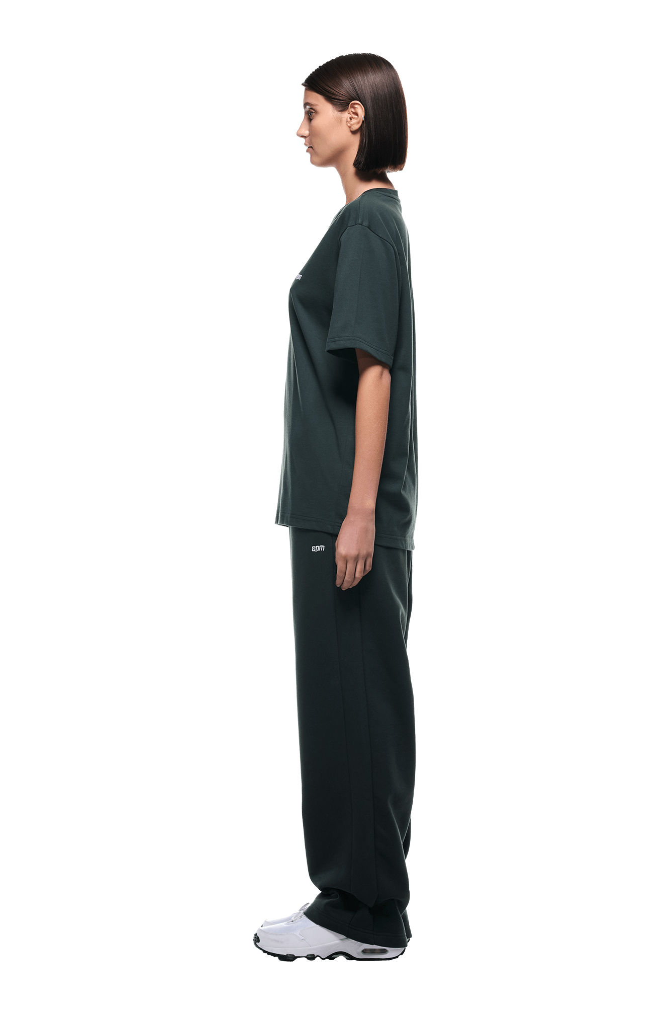 A woman with a straight bob stands in profile, wearing a 6PM T-SHIRT DARK EMERALD, matching wide-leg pants, and white sneakers on a transparent background.