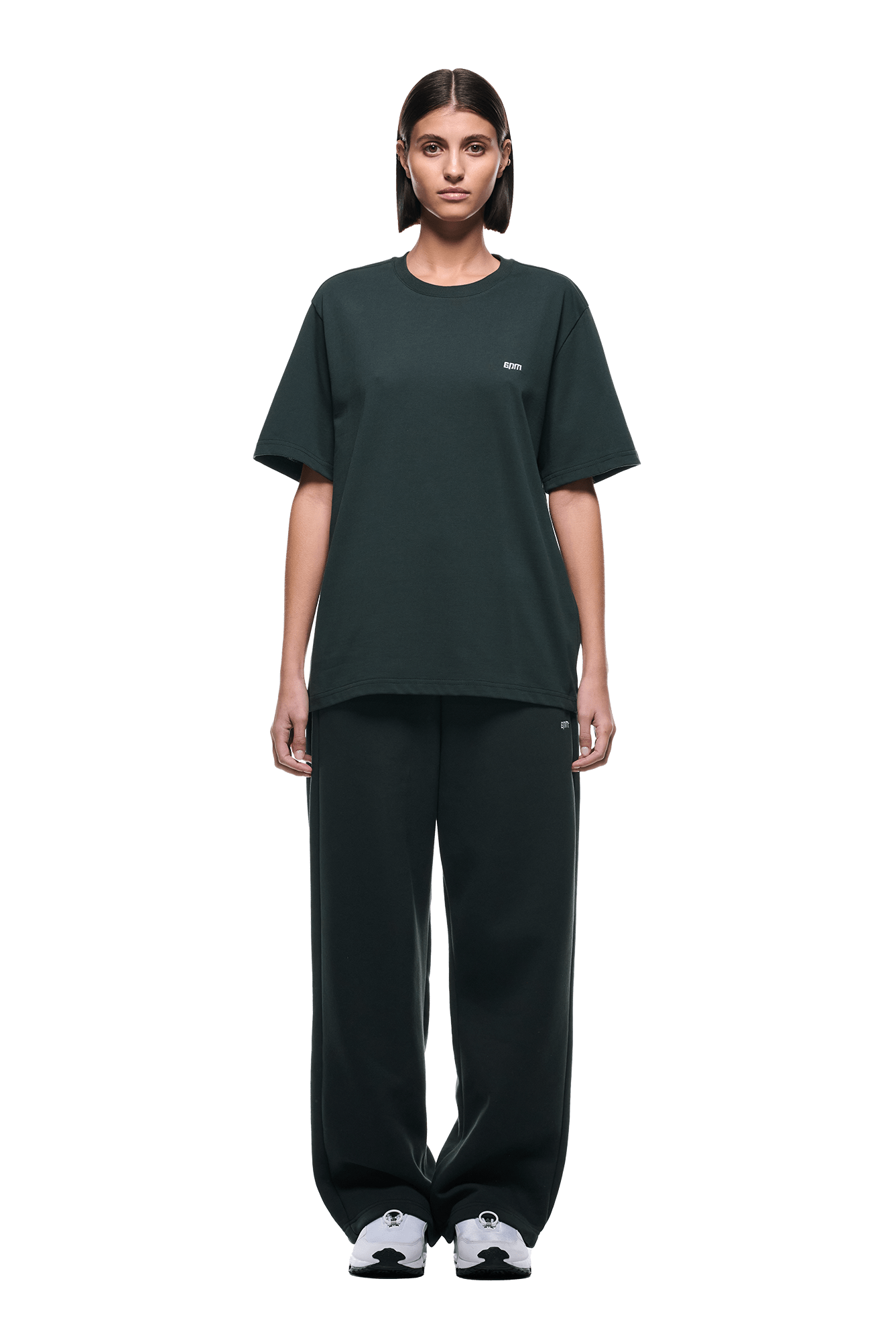 A person with short, neatly styled hair faces forward, wearing the 6PM T-SHIRT DARK EMERALD, matching loose pants, and white sneakers. The background is transparent.