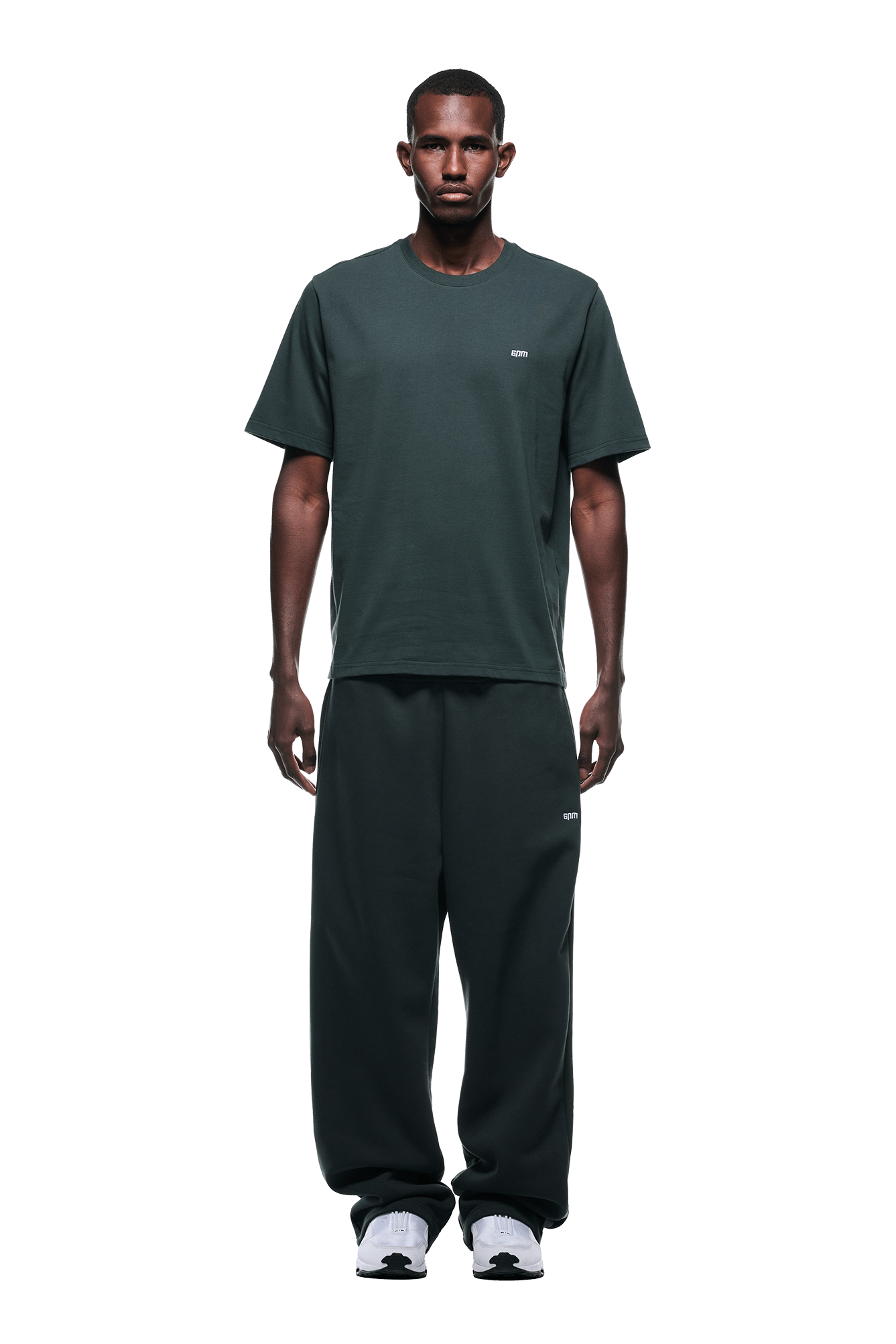 A man faces forward against a plain green background, wearing the 6PM T-SHIRT DARK EMERALD with matching baggy pants and white sneakers.