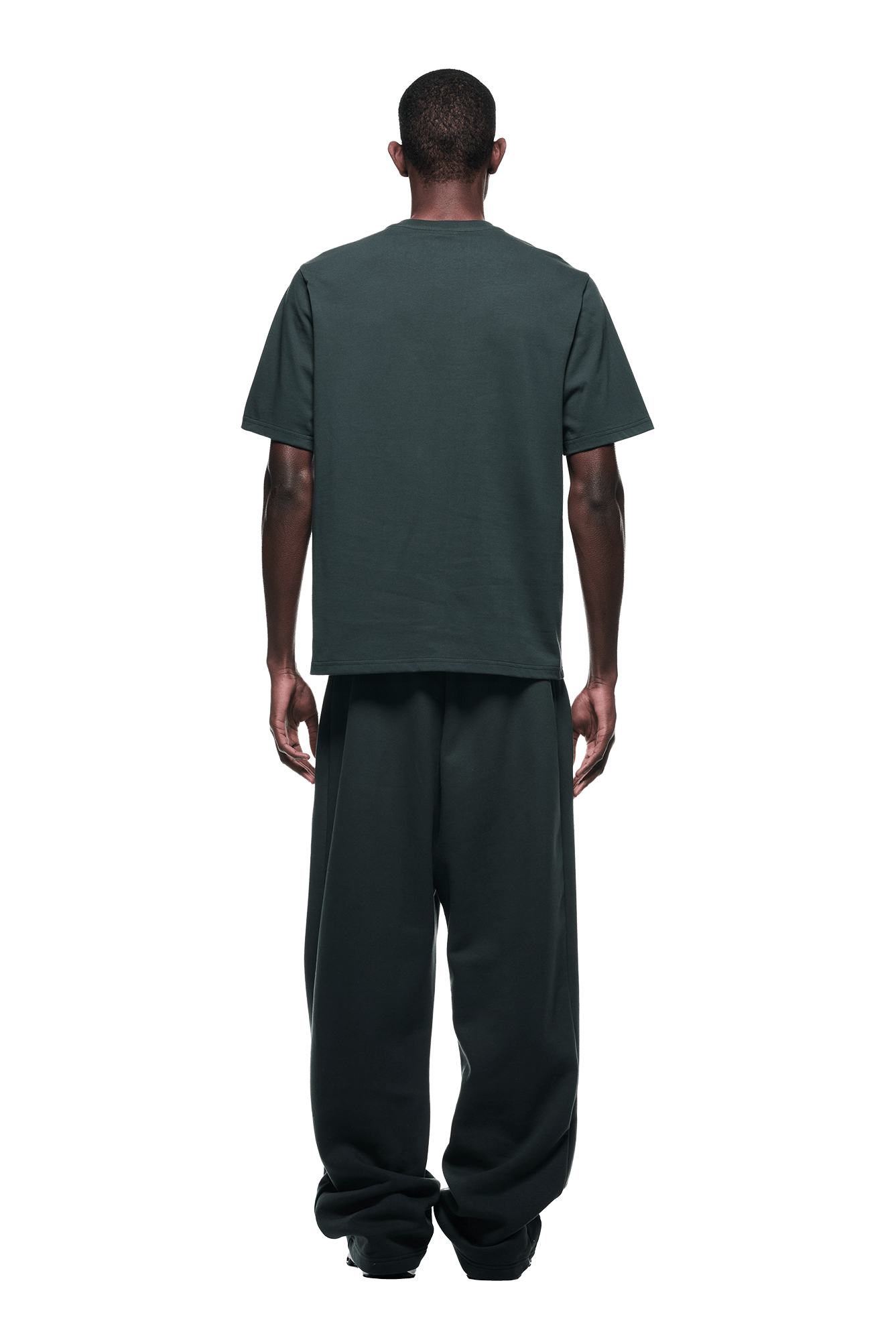 A man faces away from the camera in a loose-fitting 6PM T-SHIRT DARK EMERALD with matching wide-leg pants, arms resting by his sides against a plain background.