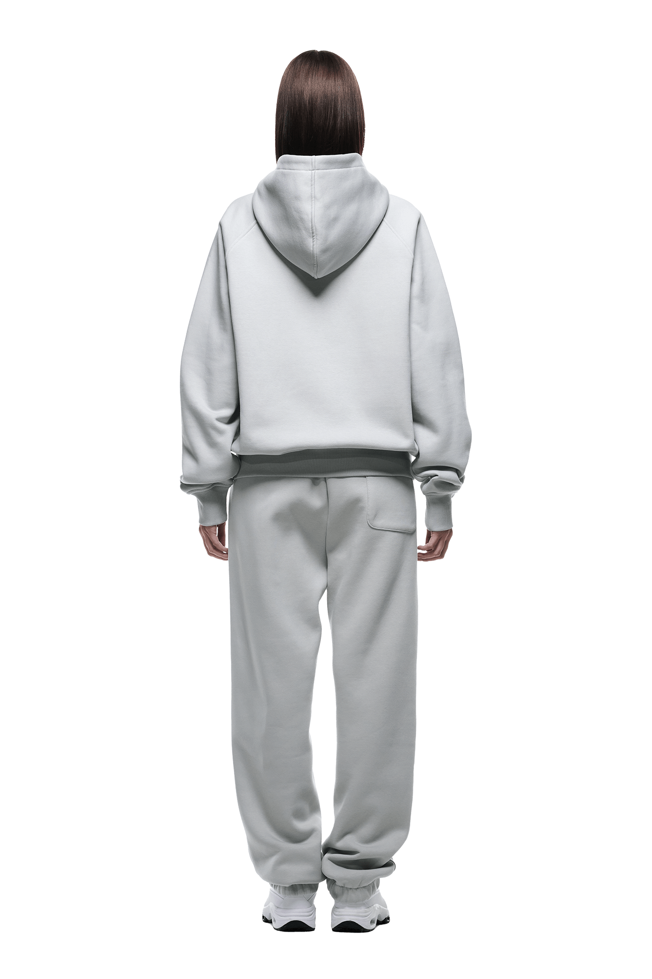 A person with straight brown hair, facing away, wears the 6PM ZIP HOODIE SMOKE GREY and matching sweatpants with white sneakers. The set’s clean look makes it perfect for daily wear. Background is transparent.