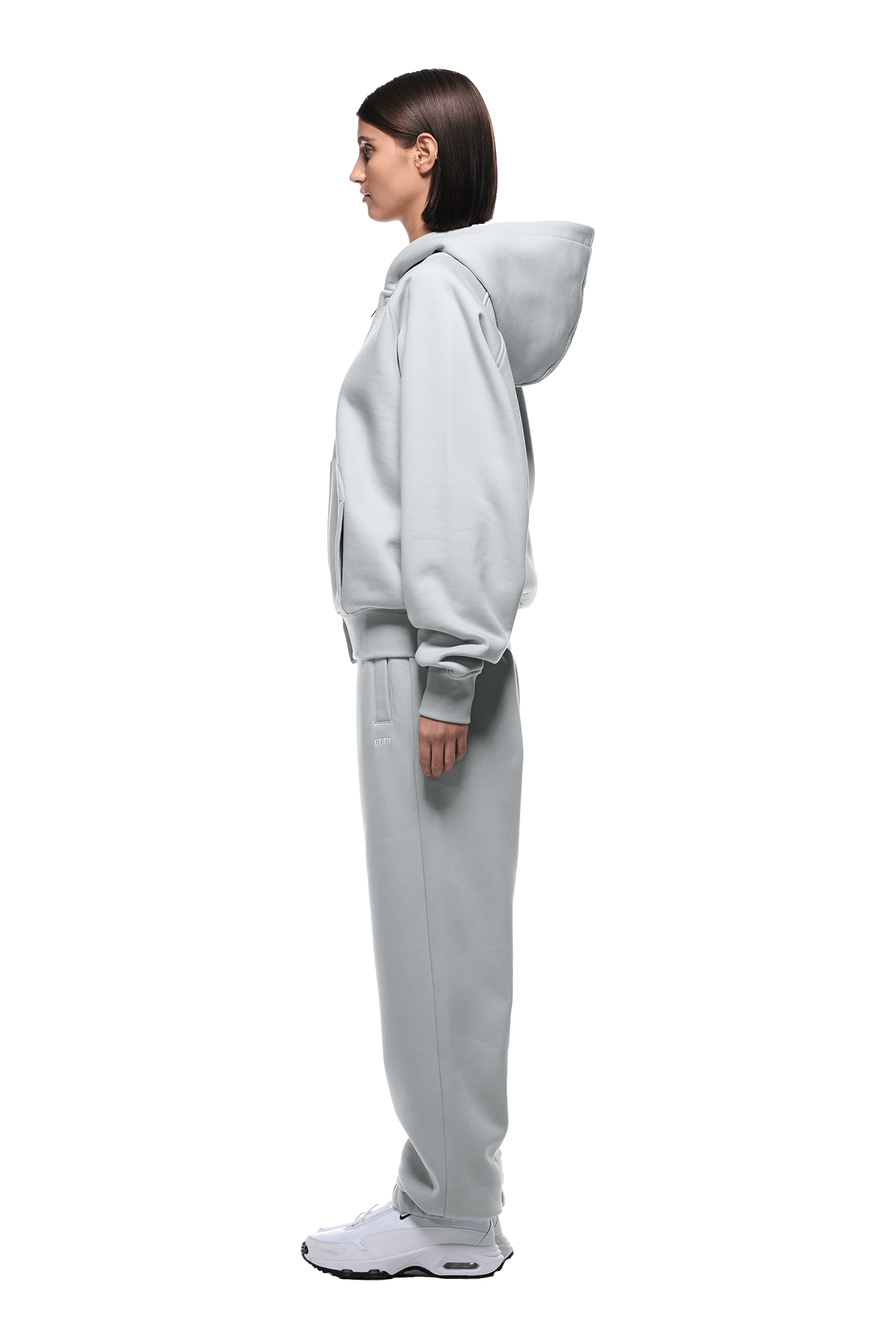 A woman in profile wears the 6PM ZIP HOODIE SMOKE GREY with matching sweatpants and white sneakers—a clean, everyday look. Her straight, chin-length hair stands out against the transparent background.