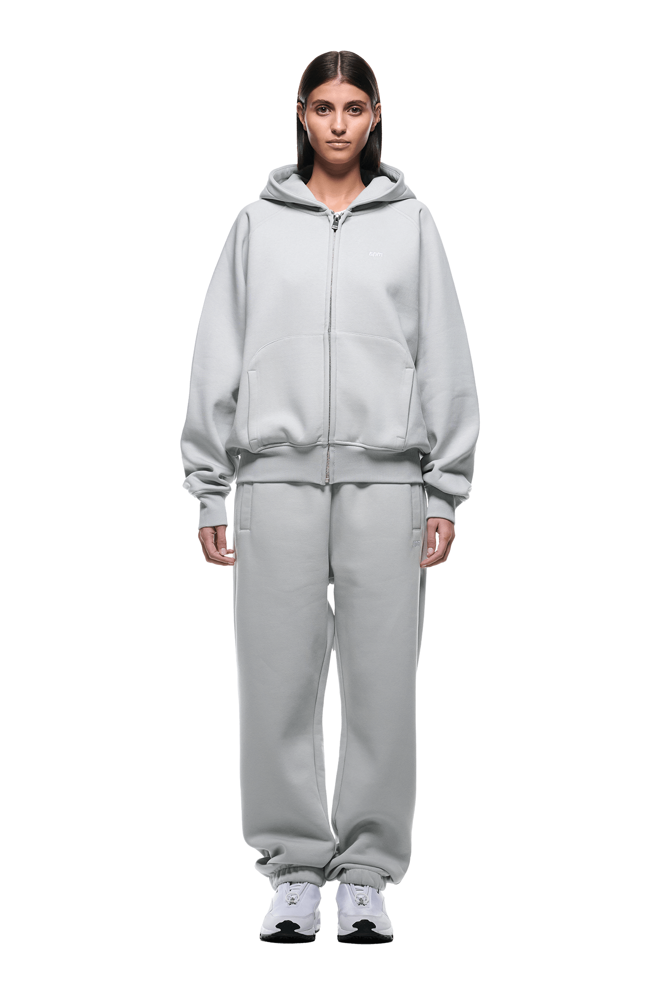 A woman faces forward wearing the 6PM ZIP HOODIE SMOKE GREY and matching sweatpants with white sneakers. Her straight hair is tucked behind her ears. The background is transparent.