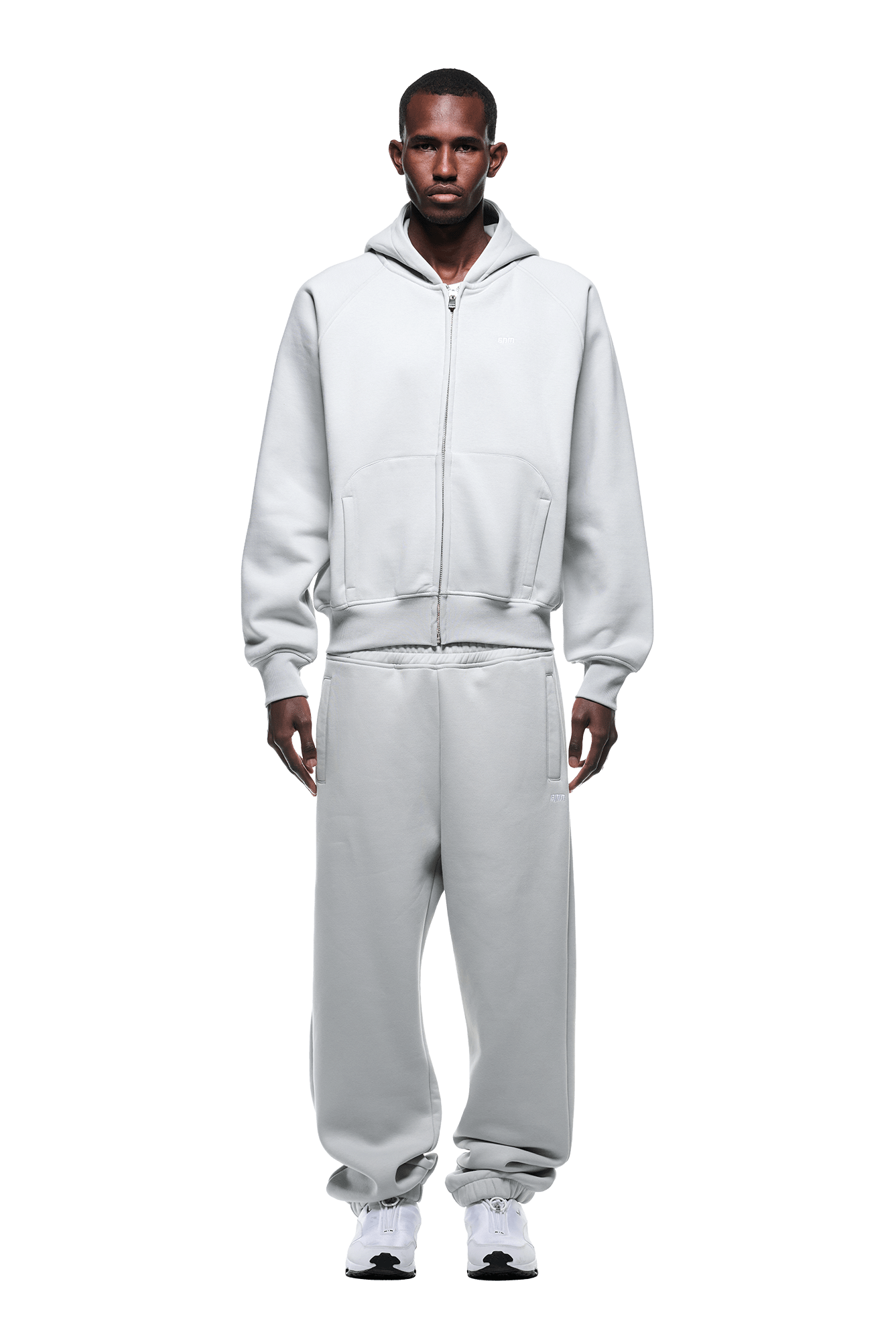 A man stands facing forward in the 6PM ZIP HOODIE SMOKE GREY set with white sneakers. The outfit has a clean look, ideal for daily wear. Background is transparent.
