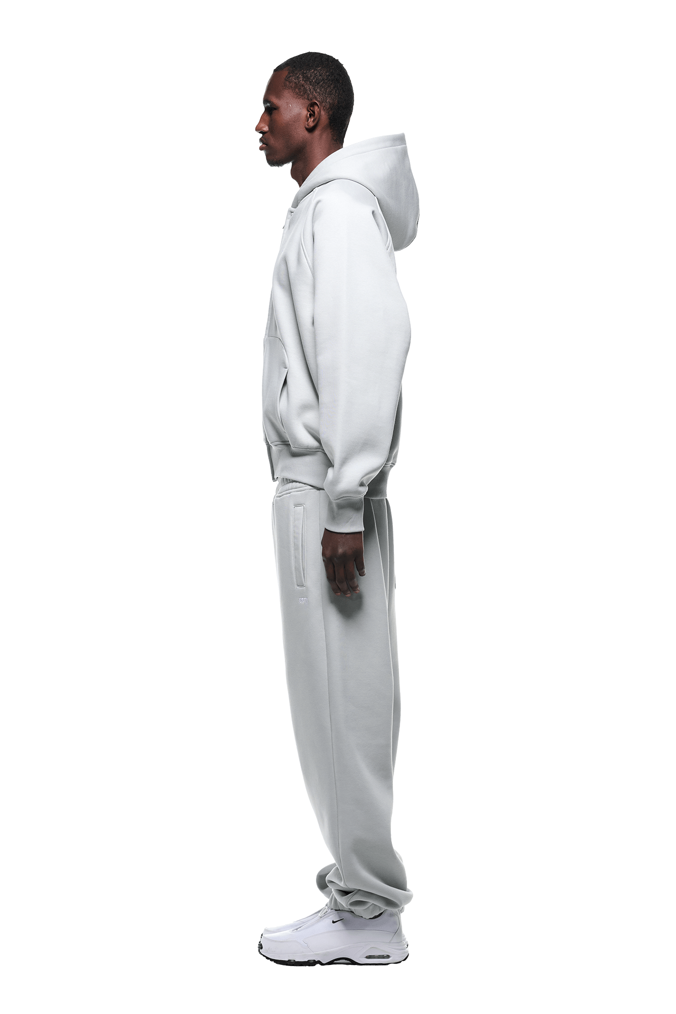 A man in profile faces left, wearing the 6PM ZIP HOODIE SMOKE GREY and matching sweatpants, paired with white sneakers against a plain green background.