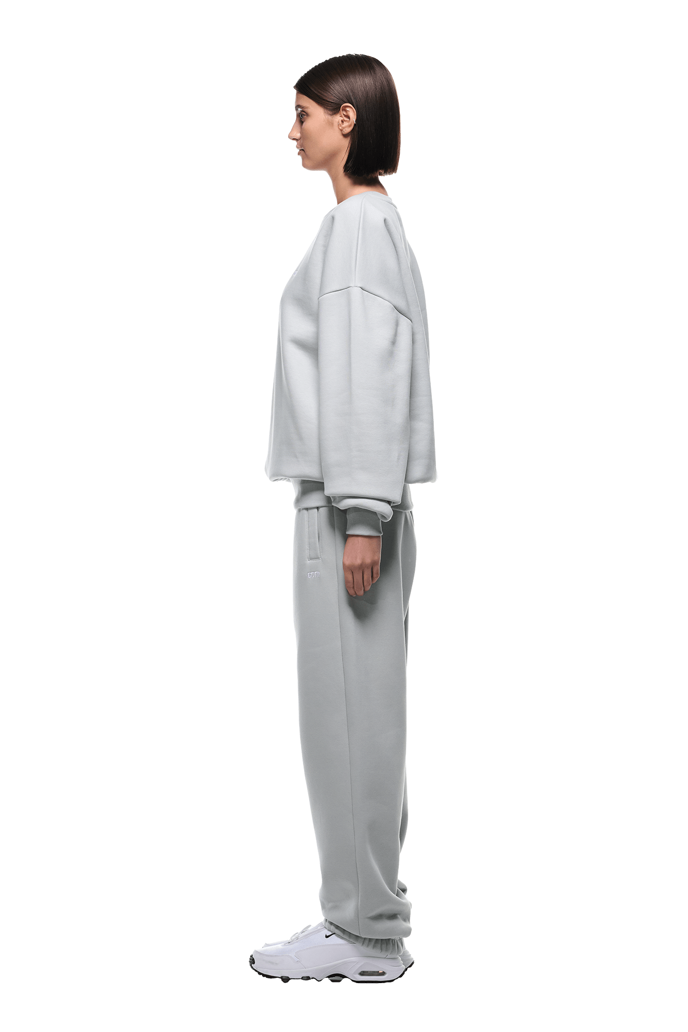 A woman with a straight bob faces left in profile, wearing the 6PM CREWNECK SMOKE GREY oversized sweatshirt with ribbed trims, matching sweatpants, and white sneakers. The background is transparent.