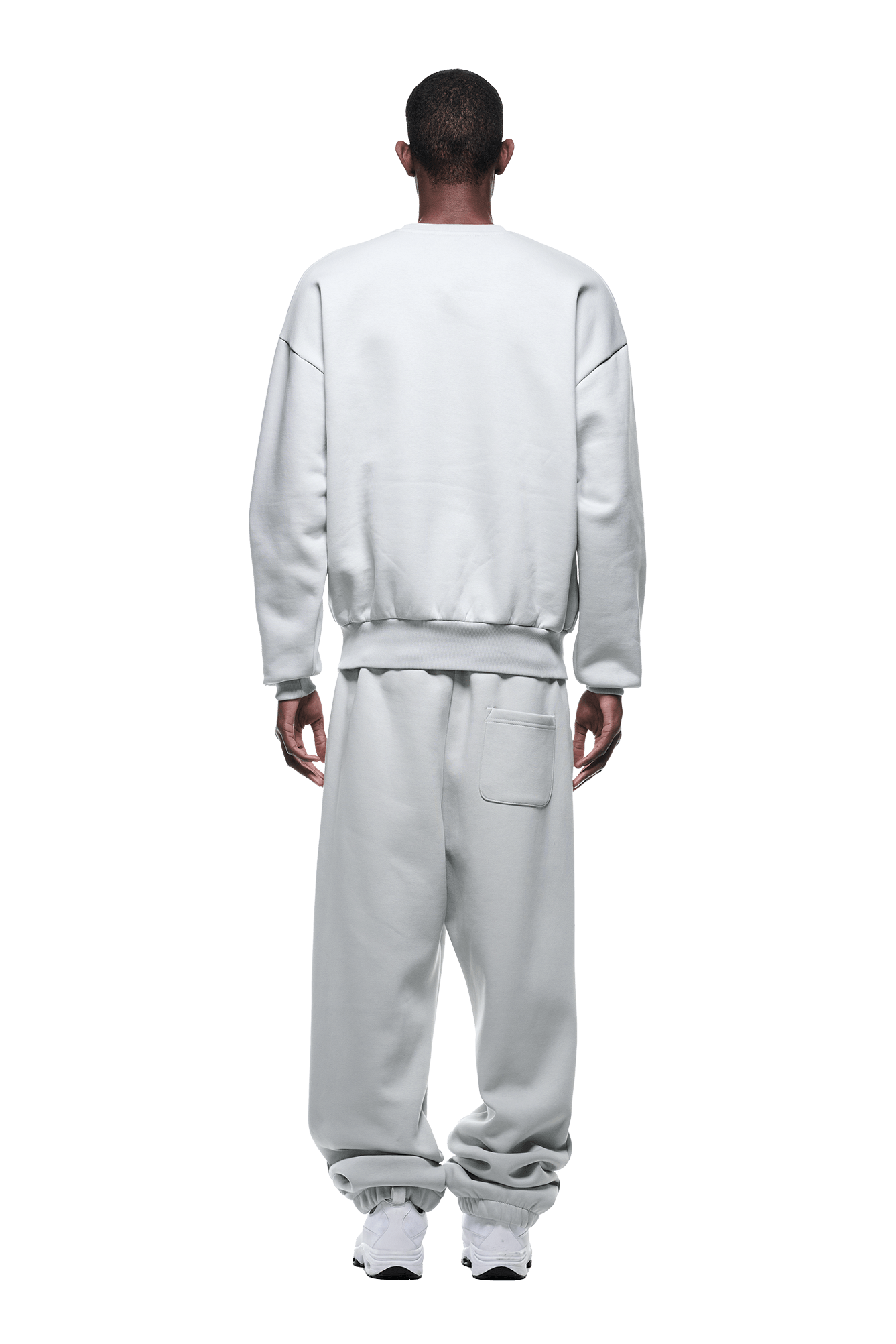A person stands with their back turned, wearing the 6PM CREWNECK SMOKE GREY sweatshirt and matching pants with white sneakers, set against a plain green background.