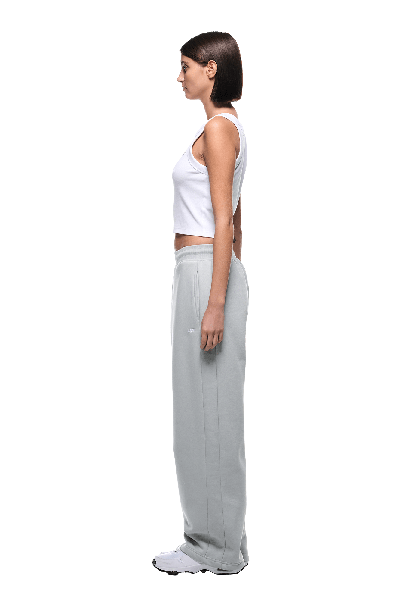 A woman with straight dark hair stands in profile, wearing a white sleeveless top, OPEN LEG JOGGER SMOKE GREY pants, and white sneakers against a plain background.
