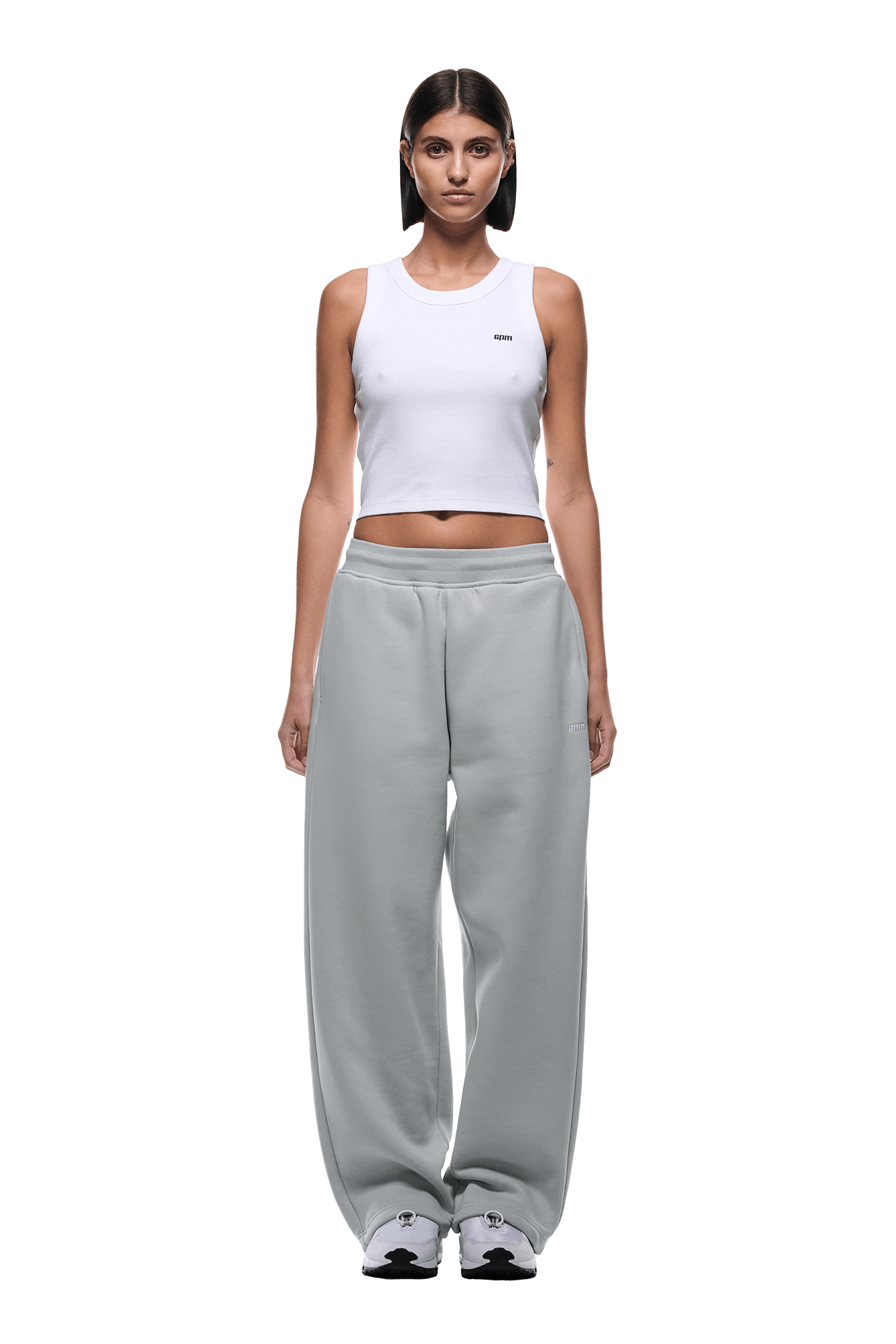 A woman with straight dark hair wears a white sleeveless crop top, OPEN LEG JOGGER SMOKE GREY sweatpants with stacking length, and white sneakers. She stands facing forward with a neutral expression on a transparent background.