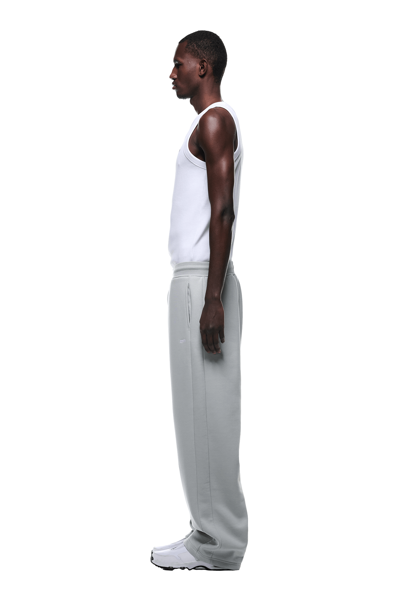 A man in profile facing left wears a white tank top, OPEN LEG JOGGER SMOKE GREY sweatpants, and white shoes against a plain background.