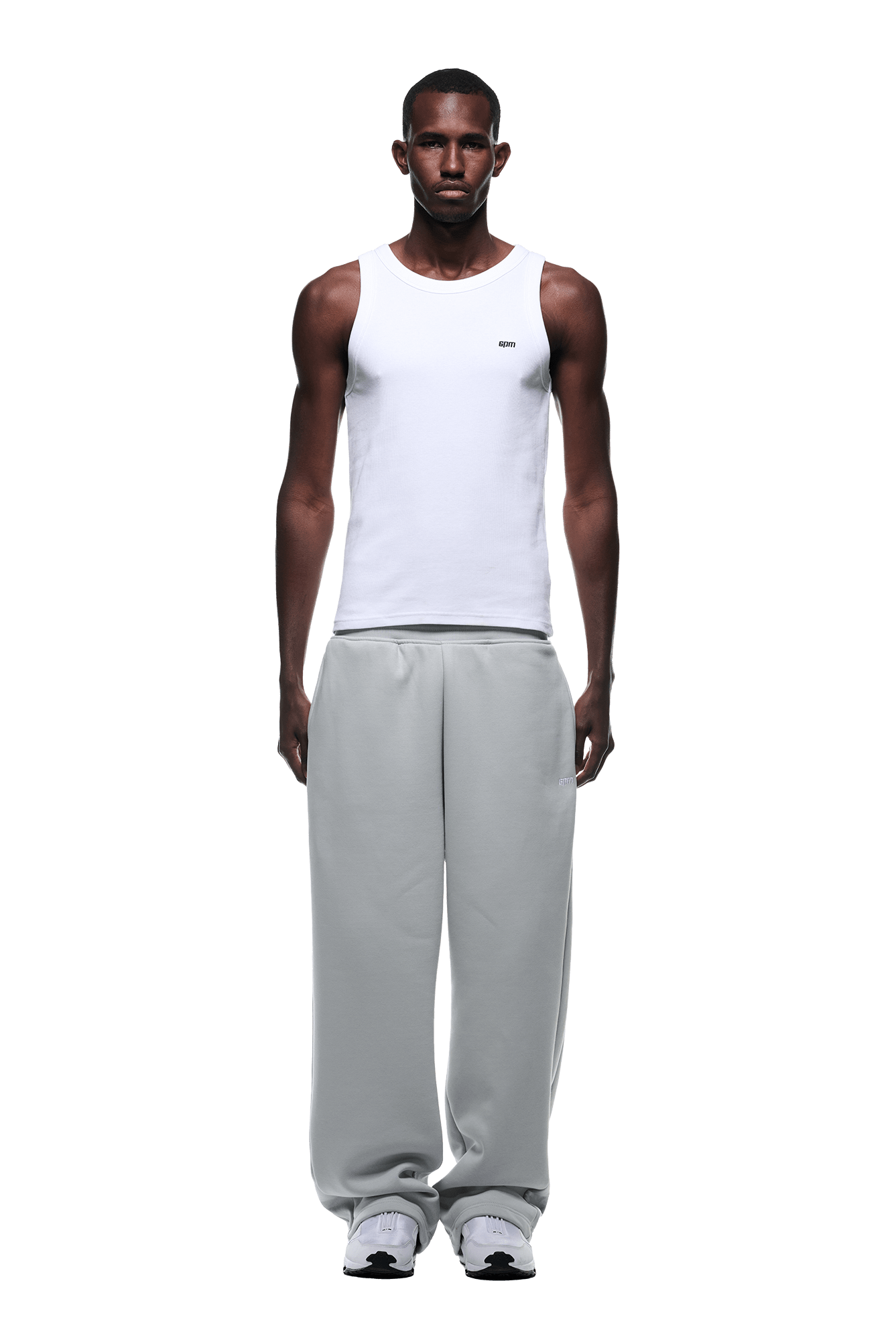 A man faces forward in a white tank top, OPEN LEG JOGGER SMOKE GREY with stacking length, and white sneakers, styled against a plain green background for a modern classic look.