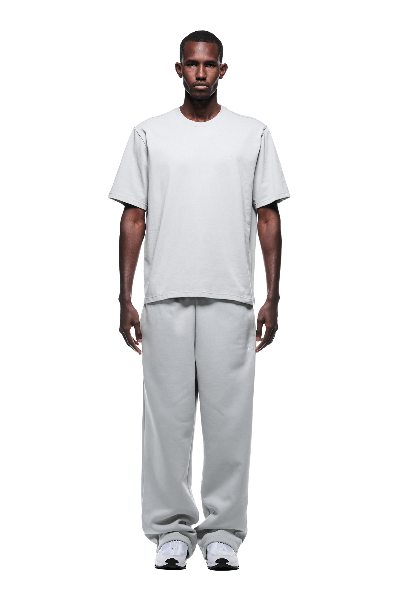 A man faces forward against a plain green background, wearing the 6PM T-SHIRT SMOKE GREY with matching wide-leg sweatpants and white sneakers.
