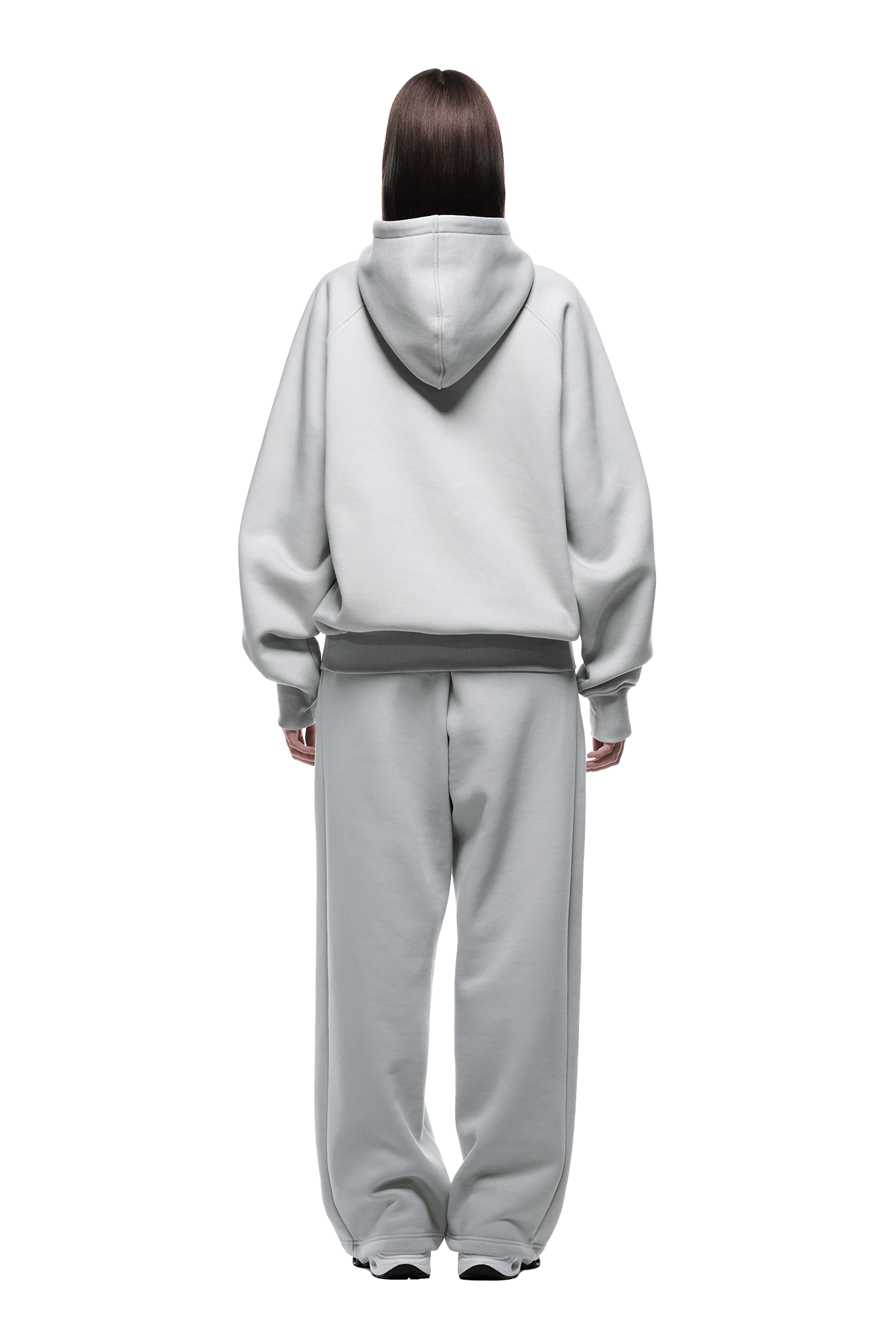 Person stands with their back to the camera, wearing the HOODIE SMOKE GREY—a light gray oversized raglan-sleeved hoodie with hood up and matching loose sweatpants. Relaxed pose, partially visible shoes, and a transparent background. Ideal for everyday wear.