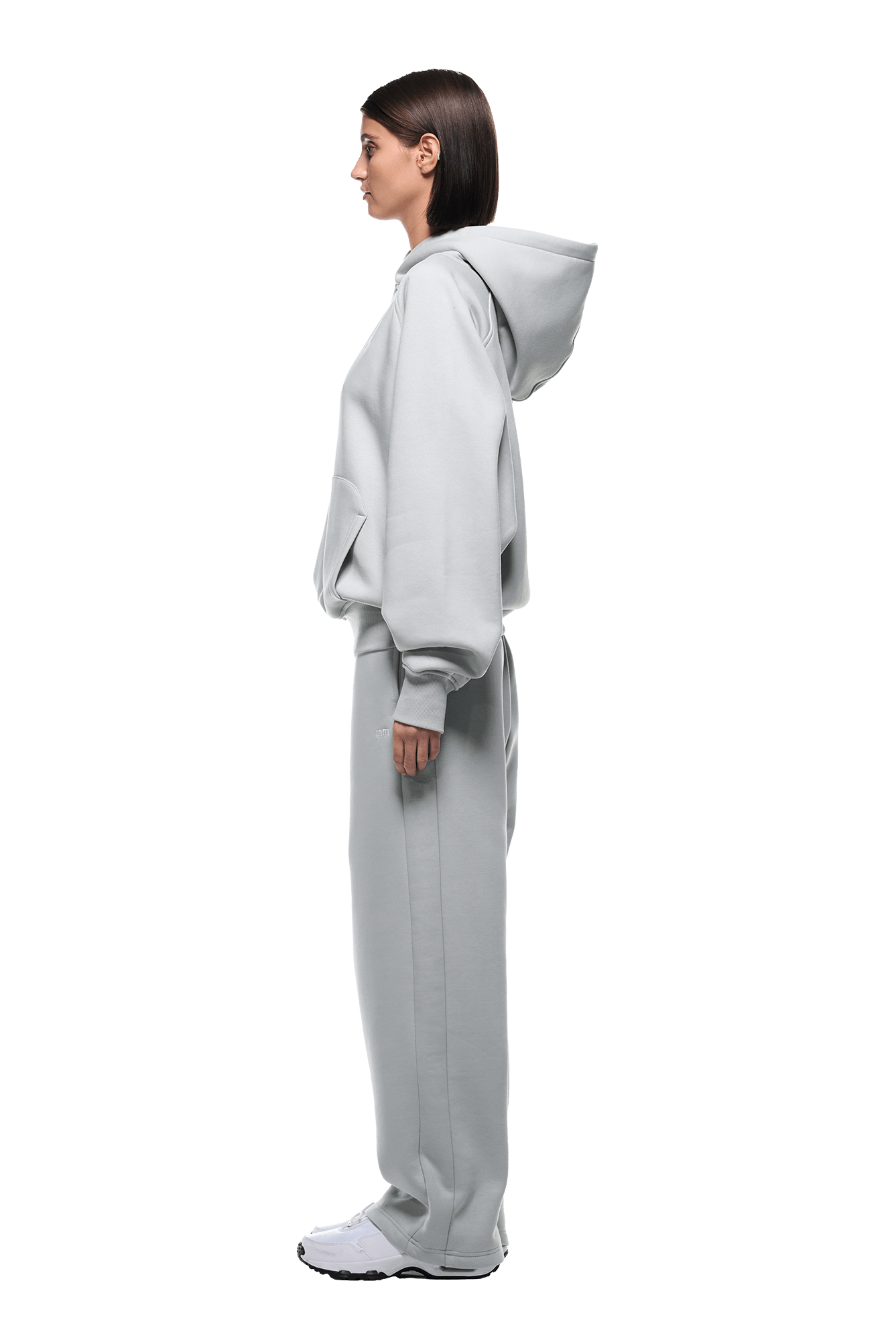 A woman in profile faces left, wearing the HOODIE SMOKE GREY with raglan sleeves and a kangaroo pocket, matching sweatpants, and white sneakers against a transparent background.