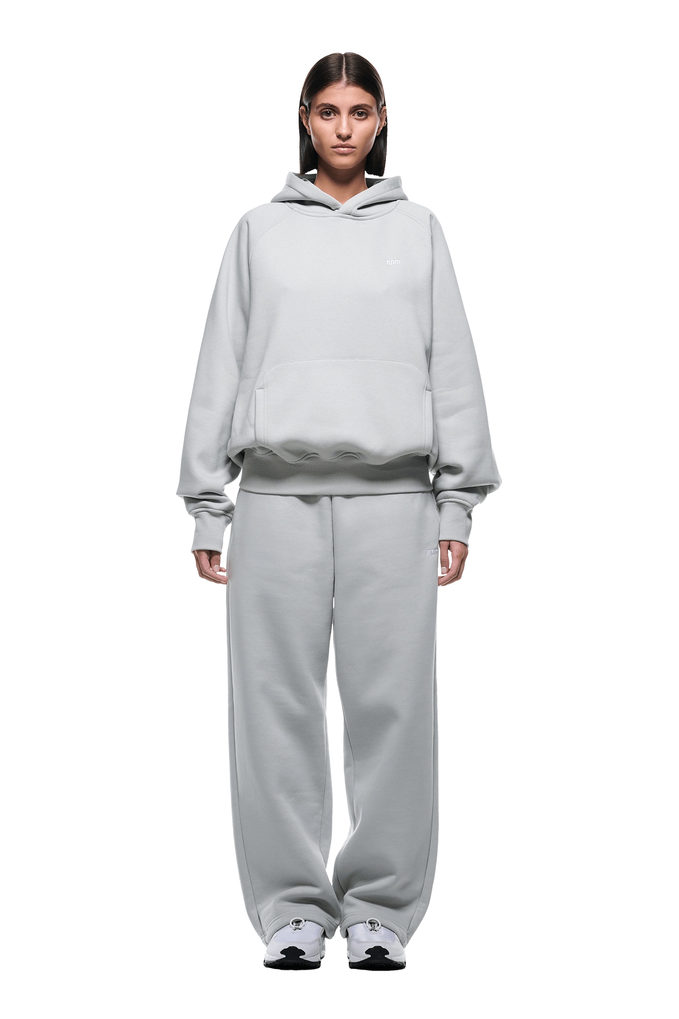 A person stands facing forward in the HOODIE SMOKE GREY with raglan sleeves, a kangaroo pocket, and matching sweatpants paired with white sneakers, perfect for daily wear, against a plain green background.