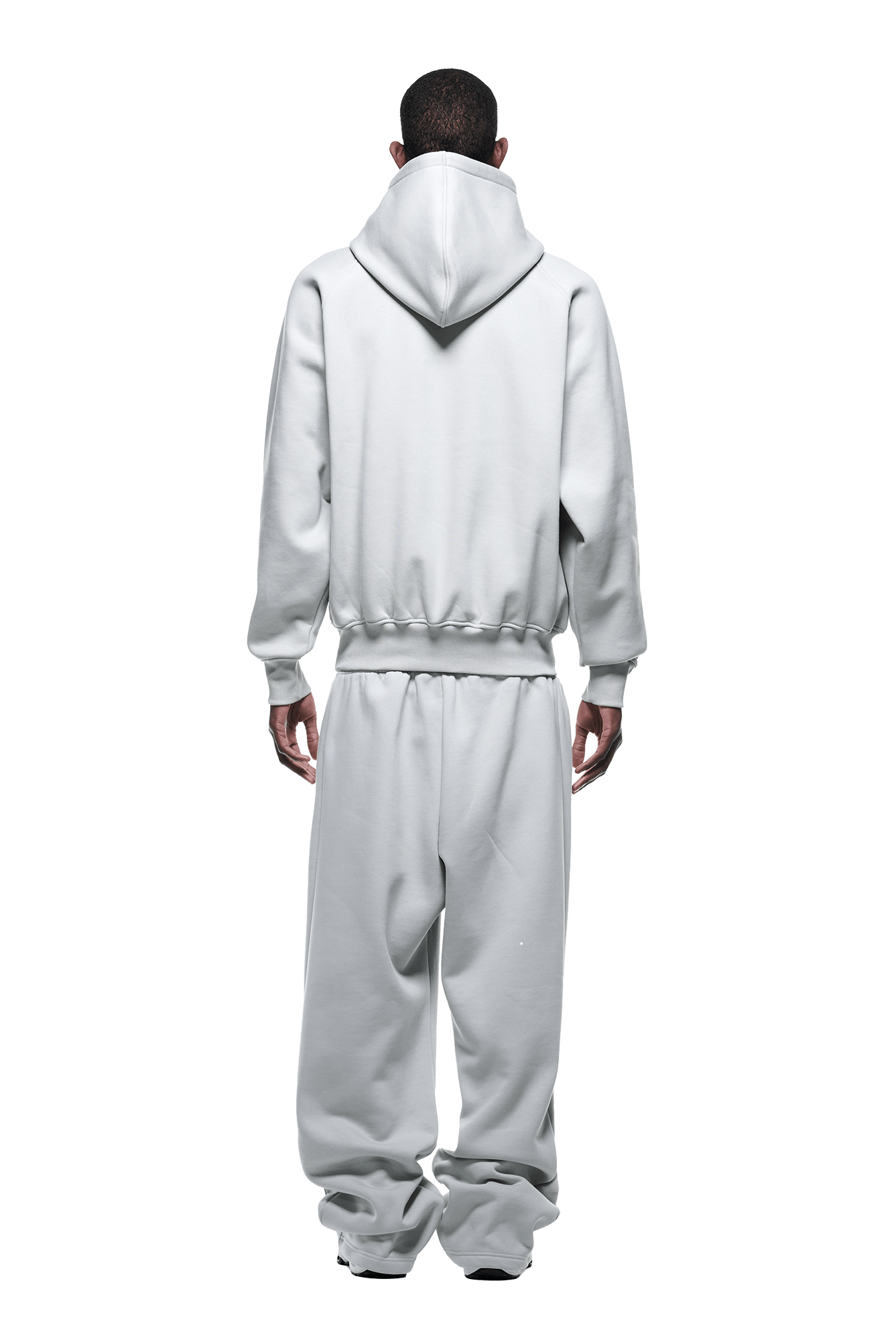 A person stands facing away, wearing the HOODIE SMOKE GREY—a loose-fitting light gray raglan-sleeved hoodie with matching sweatpants. The hood is down, hands at their sides. Ideal for everyday use. Background is transparent.