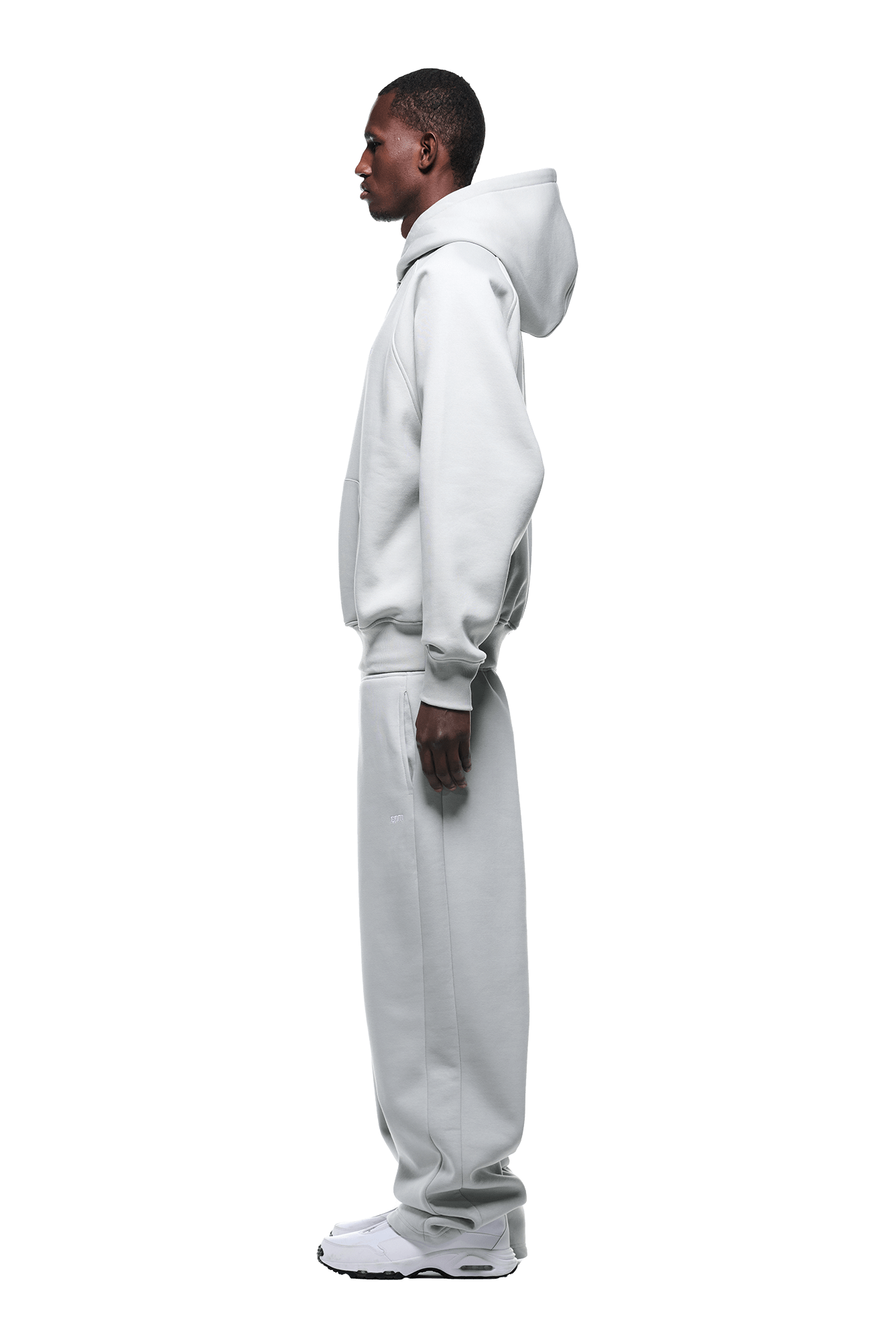 A person stands in profile wearing the HOODIE SMOKE GREY, a loose-fitting, light gray raglan-sleeved hoodie with a kangaroo pocket and matching sweatpants, paired with white sneakers against a plain green background.