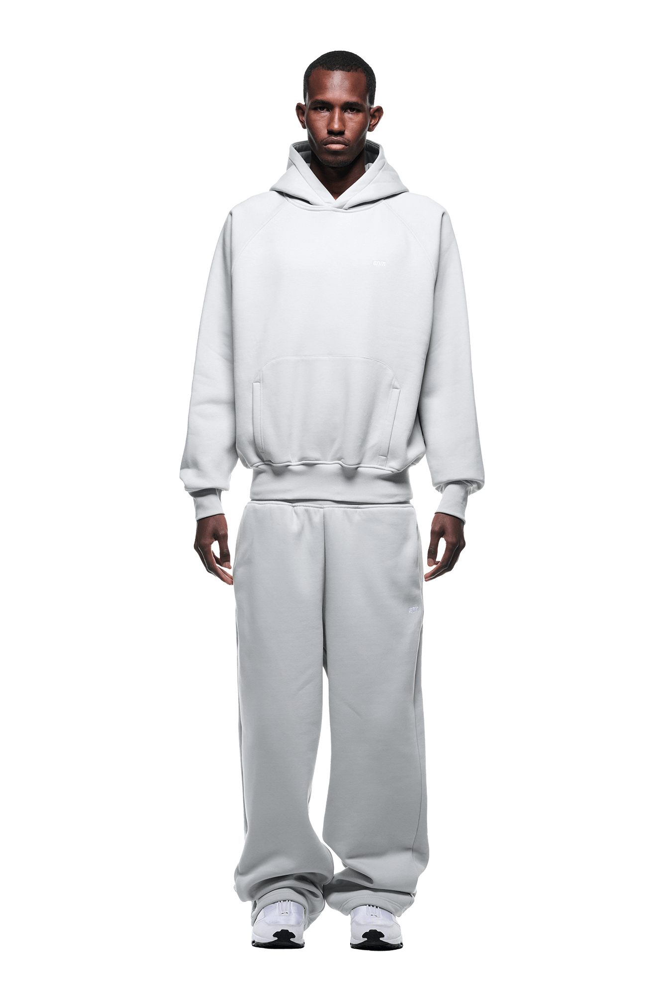 A man stands facing forward in a HOODIE SMOKE GREY with raglan sleeves and a kangaroo pocket, matching sweatpants, and white sneakers. Against a transparent background, his hands rest at his sides, highlighting this everyday outfit.