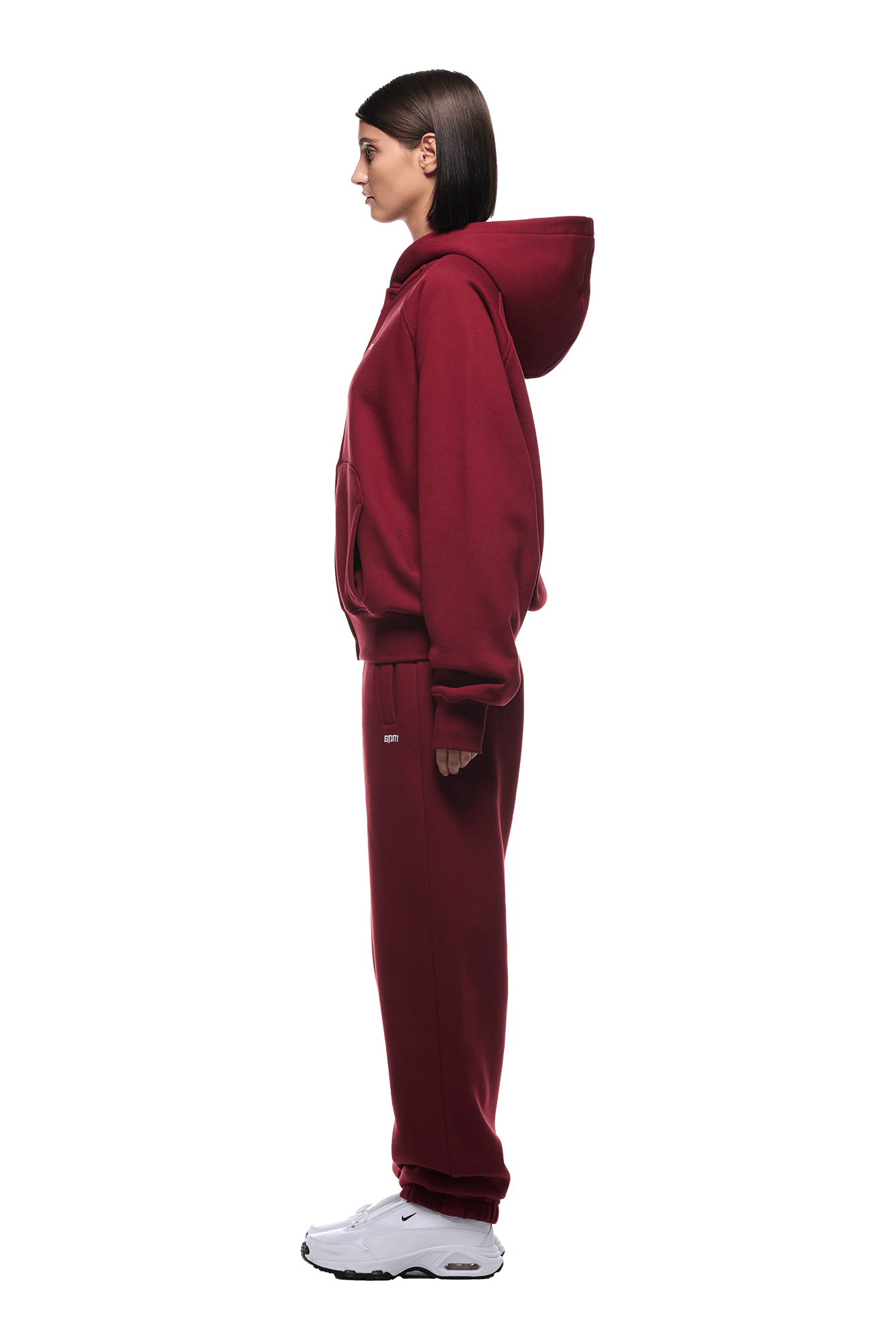 A woman in profile wears the 6PM ZIP HOODIE WINE RED set with white sneakers against a plain green background—a stylish, clean-finished ensemble ideal for everyday wear.