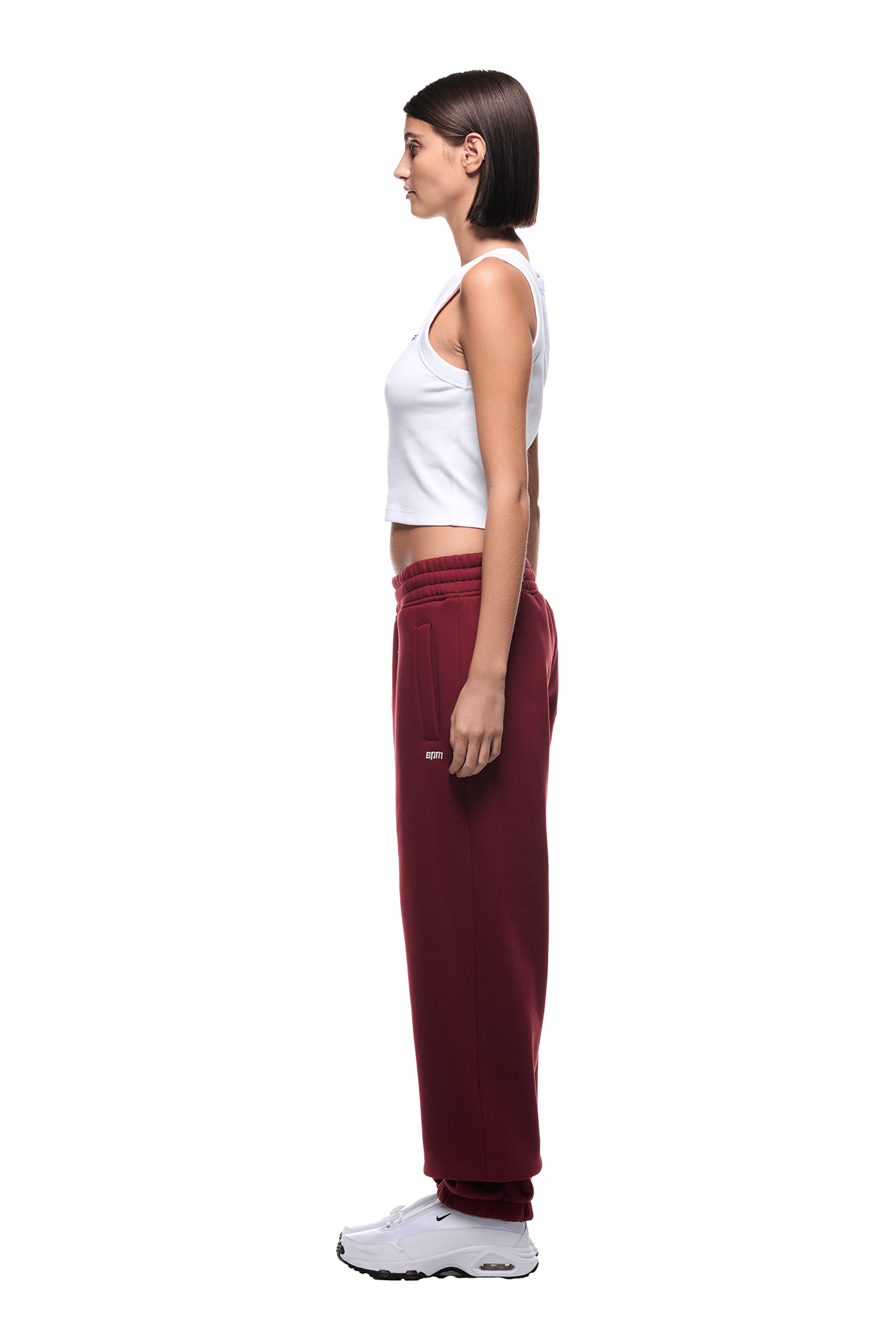 A woman with straight dark hair, in a white sleeveless crop top, 6PM CUFF JOGGER WINE RED sweatpants with cuffed legs, and white sneakers stands in profile facing left against a plain background.