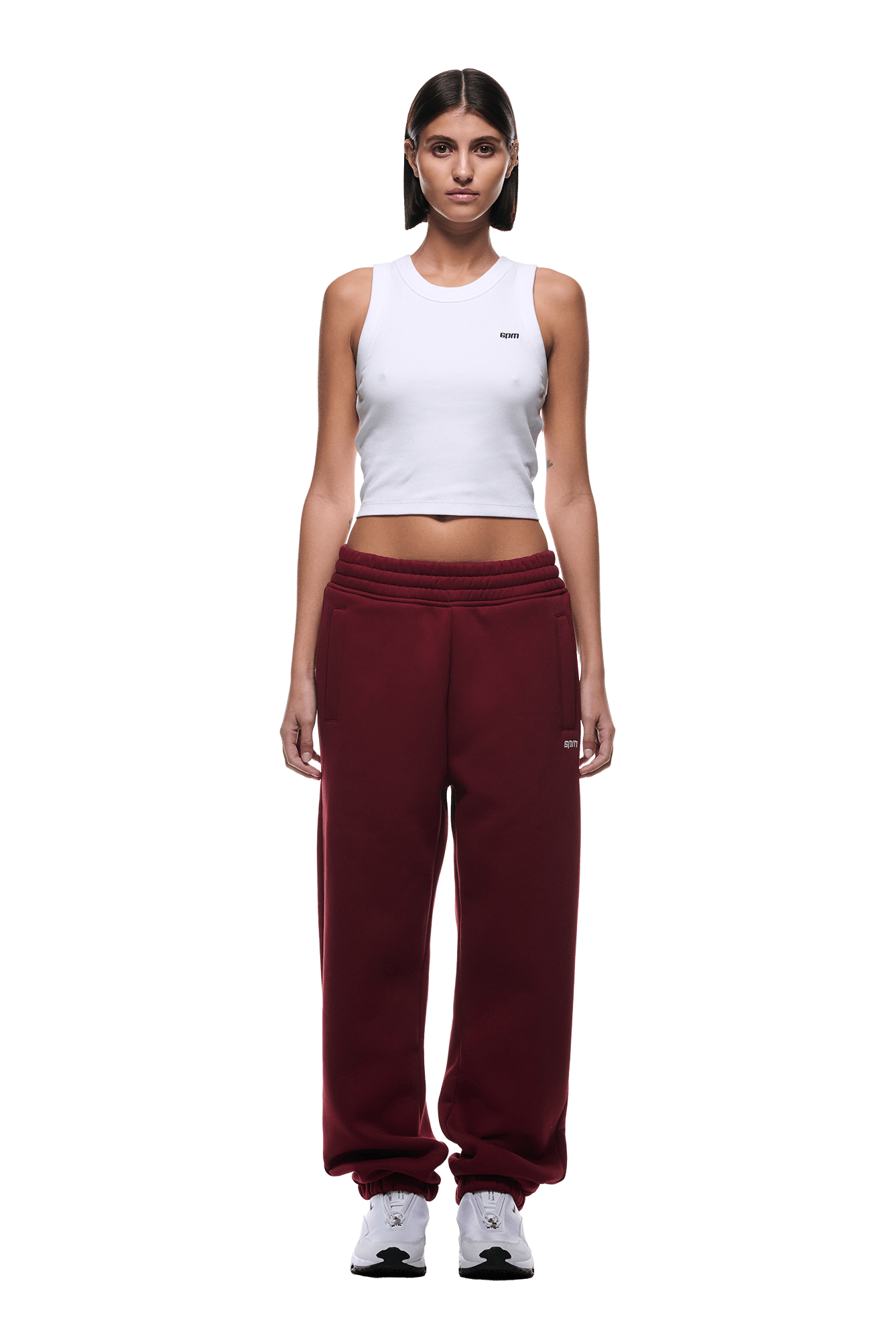 A woman with straight dark hair and a neutral expression faces forward, wearing 6PMs CUFF JOGGER WINE RED sweatpants, a white sleeveless crop top, and white sneakers. The background is transparent.