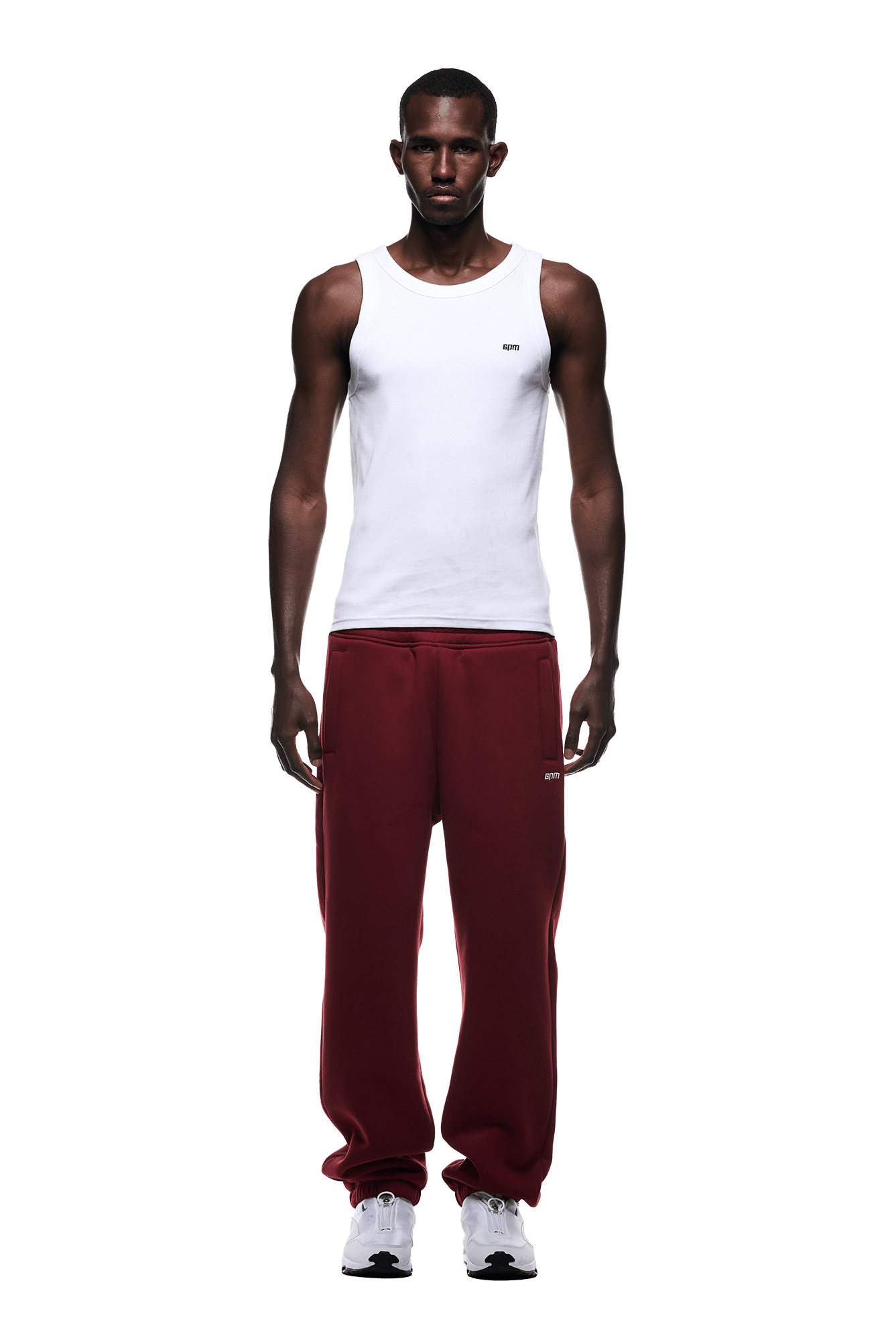 A man faces forward, wearing a white tank top, 6PM CUFF JOGGER WINE RED sweatpants with cuffed leg openings, and white sneakers. The background is transparent.