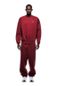 A man stands facing forward in a matching 6PM CREWNECK WINE RED oversized sweatshirt and sweatpants with small white logos, ribbed trims, and white sneakers, set against a plain white background.