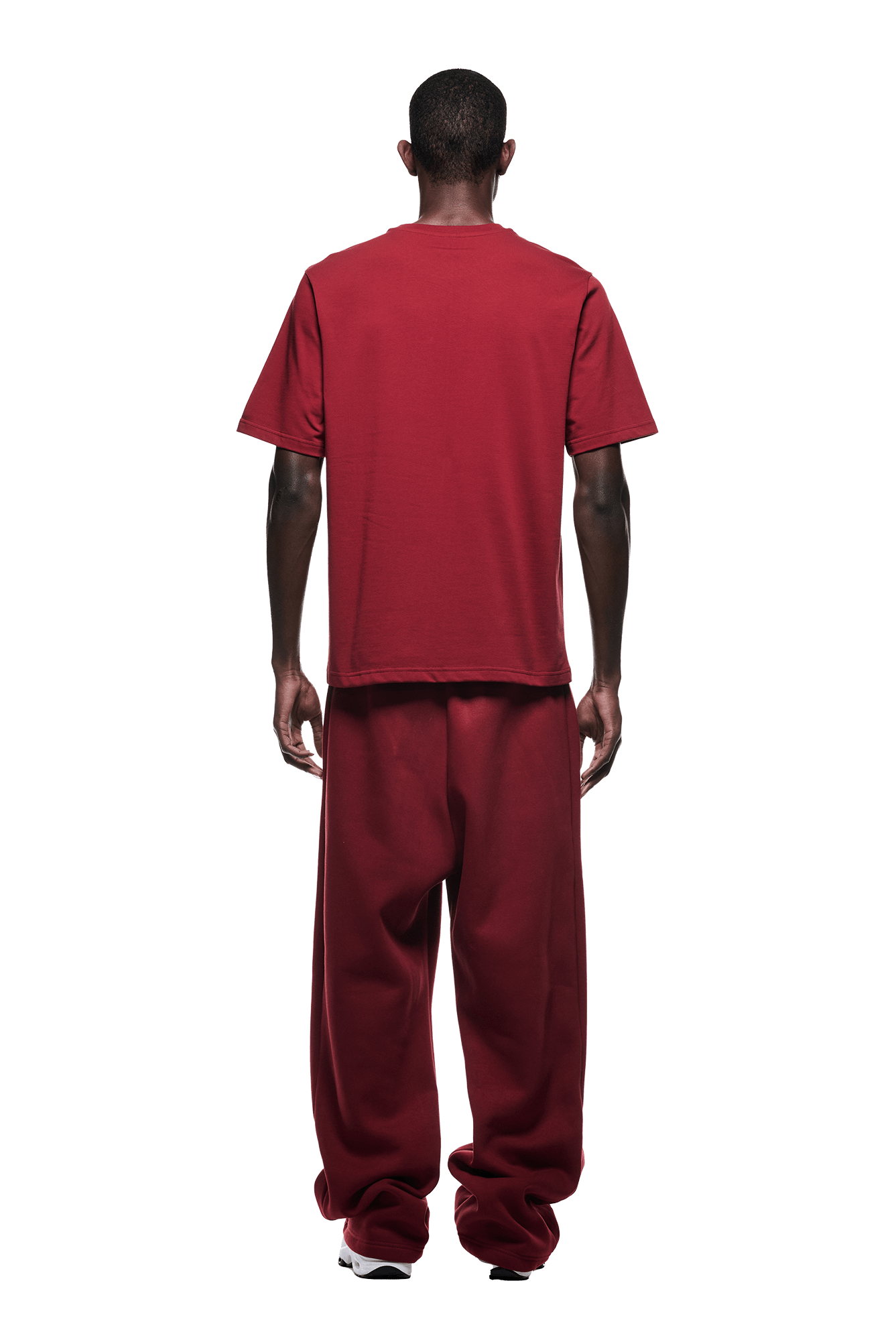 A man faces away, wearing a 6PM T-SHIRT WINE RED and red baggy pants with white sneakers, set against a plain green background.