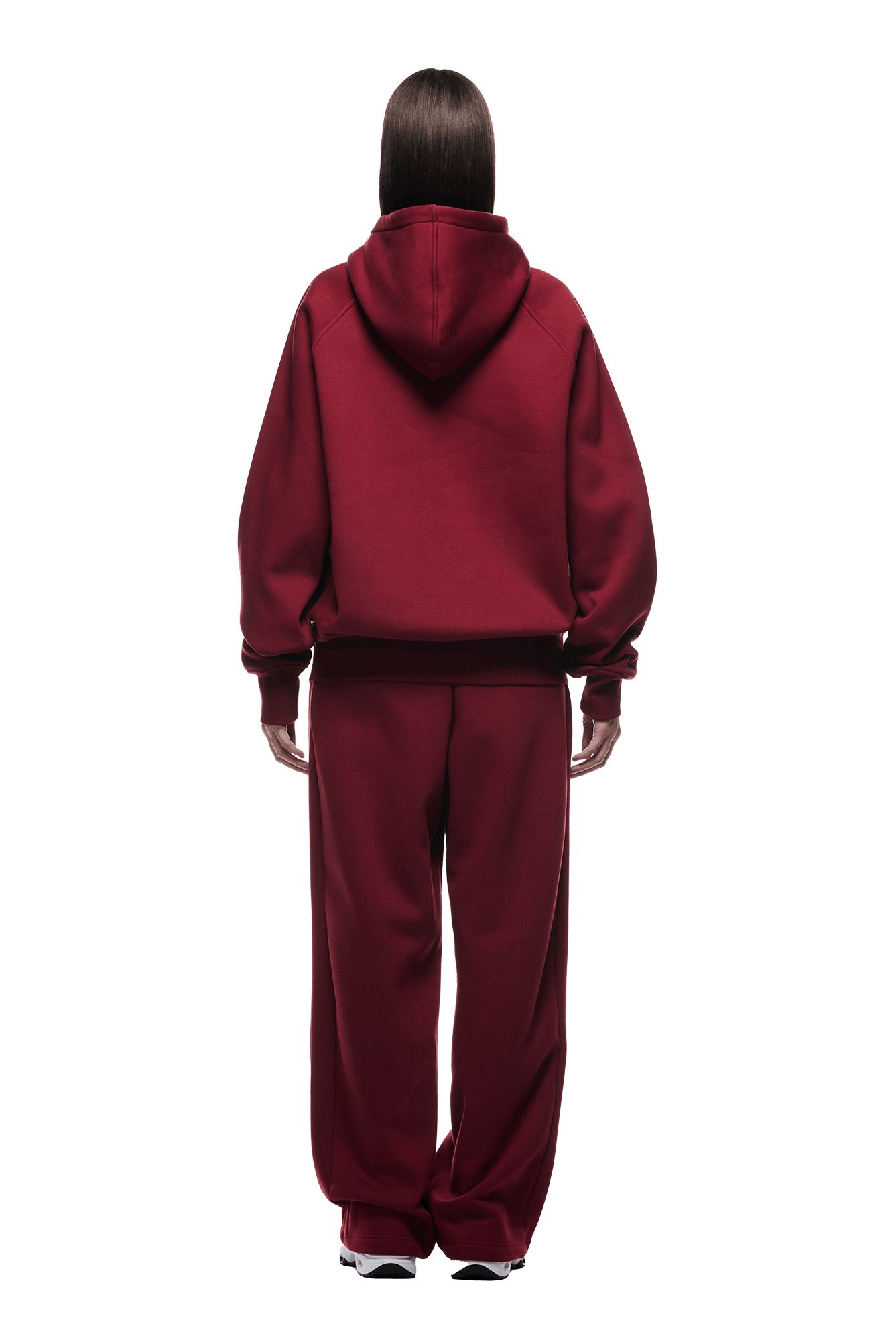 A person with straight, long brown hair faces away, wearing the HOODIE WINE RED—loose-fitting with raglan sleeves and a kangaroo pocket—paired with matching sweatpants and white sneakers against a green background.