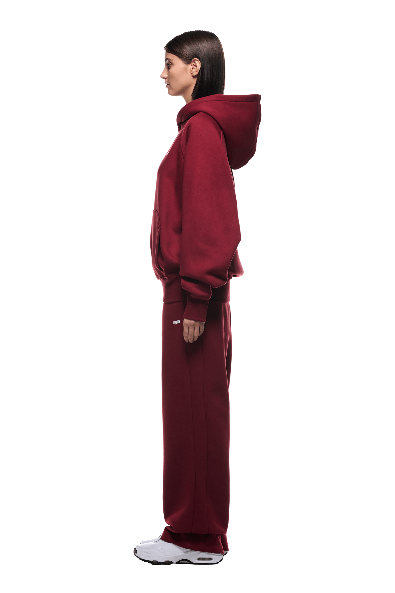 A woman in profile wears the HOODIE WINE RED with matching sweatpants and white sneakers. Her straight dark hair complements the casual, everyday look as she faces forward against a transparent background.