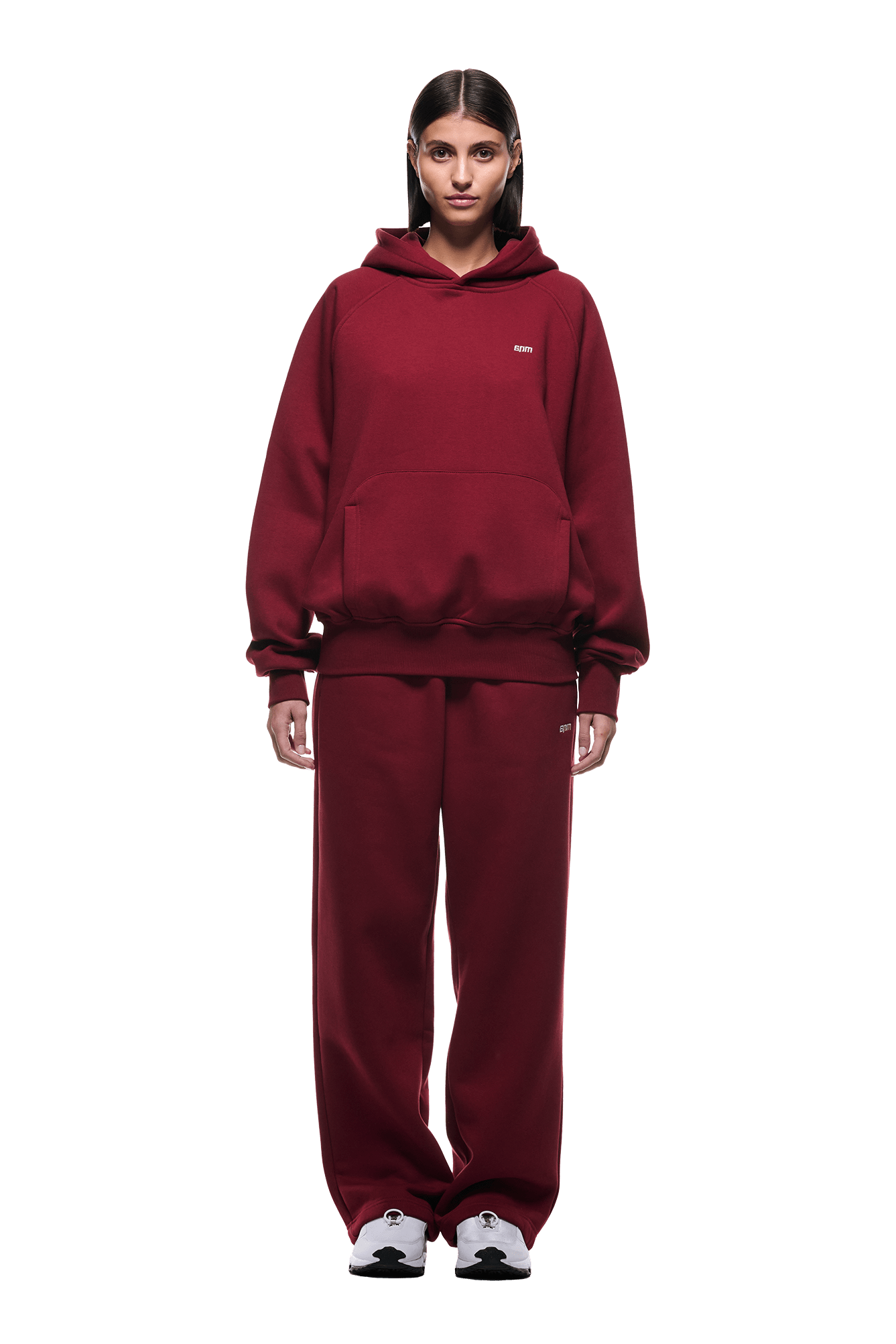 A person with straight, dark hair parted in the middle wears the loose-fitting HOODIE WINE RED and matching sweatpants, both ideal for everyday use. The hoodie features raglan sleeves and a kangaroo pocket, paired with white sneakers.