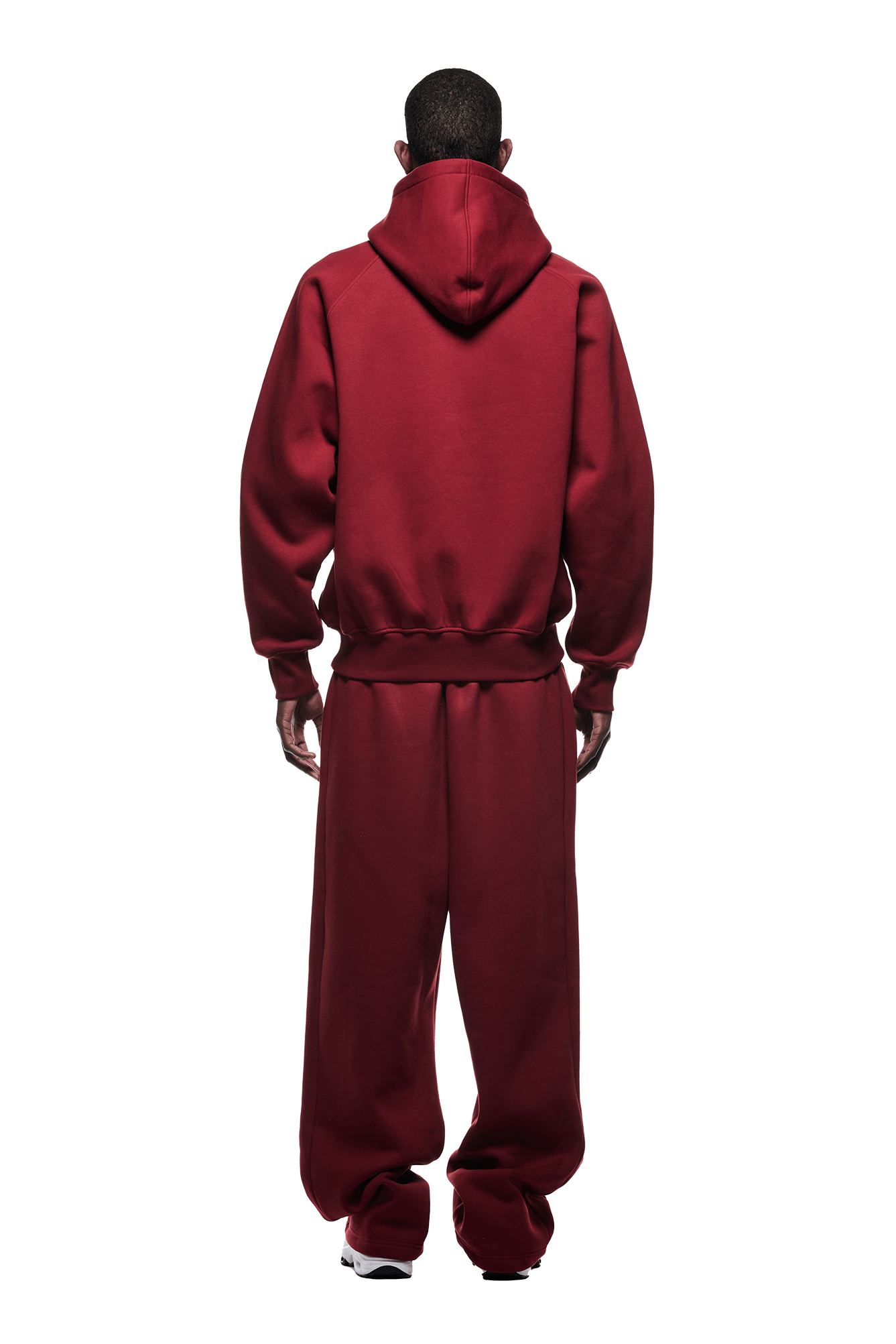 A man faces away, wearing the HOODIE WINE RED—a loose-fitting maroon raglan-sleeved hoodie with a kangaroo pocket and matching sweatpants with white sneakers—against a plain green background, ideal for everyday wear.