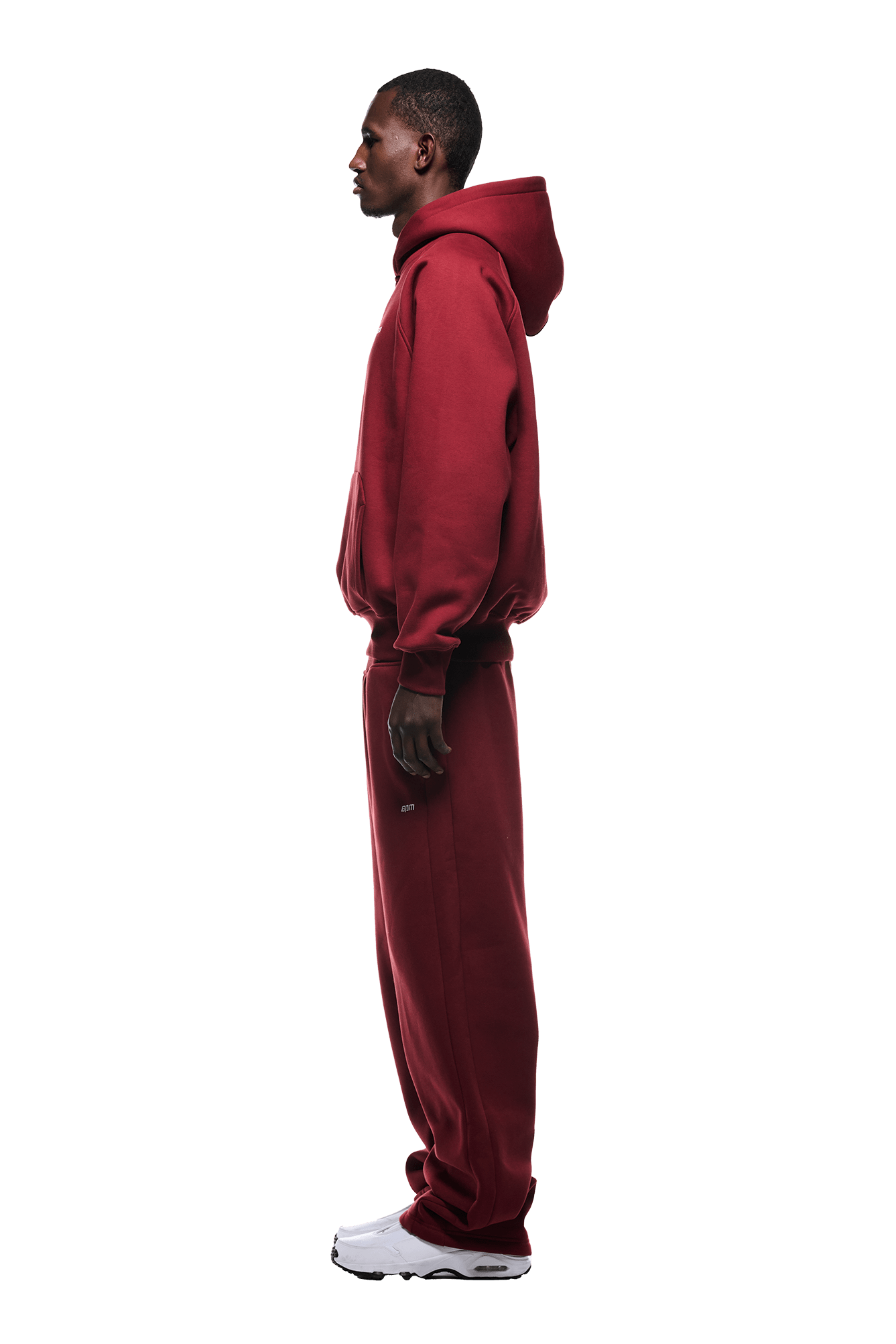 A person in profile wears the HOODIE WINE RED with raglan sleeves and a kangaroo pocket, paired with matching wine red sweatpants and white sneakers against a green background—ideal for everyday wear.
